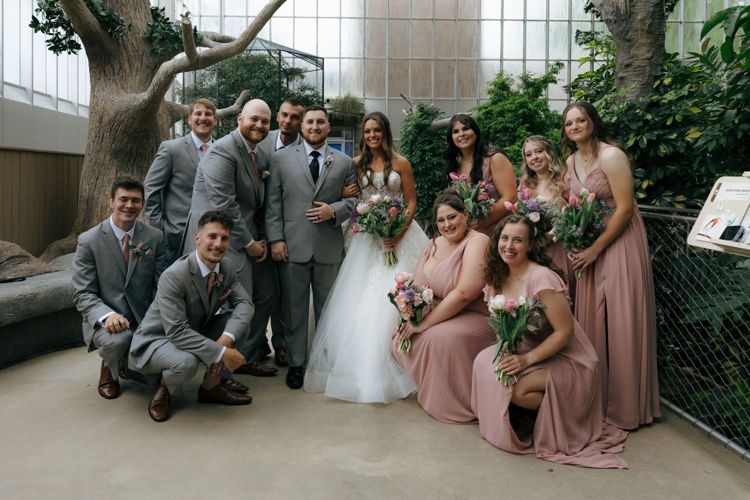 Wedding party poses indoors with bride in white gown, groomsmen in gray suits, and bridesmaids in dusty rose dresses.