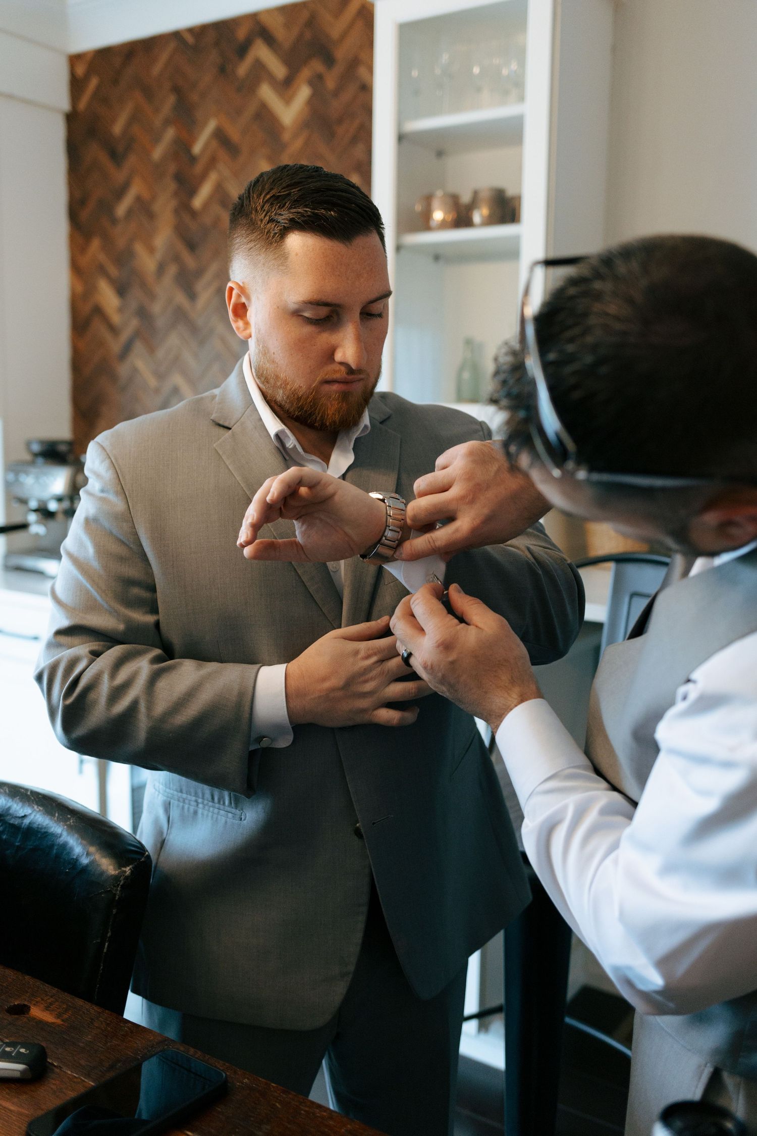 A man in a gray suit gets help adjusting his cufflinks from another man in a white shirt indoors.