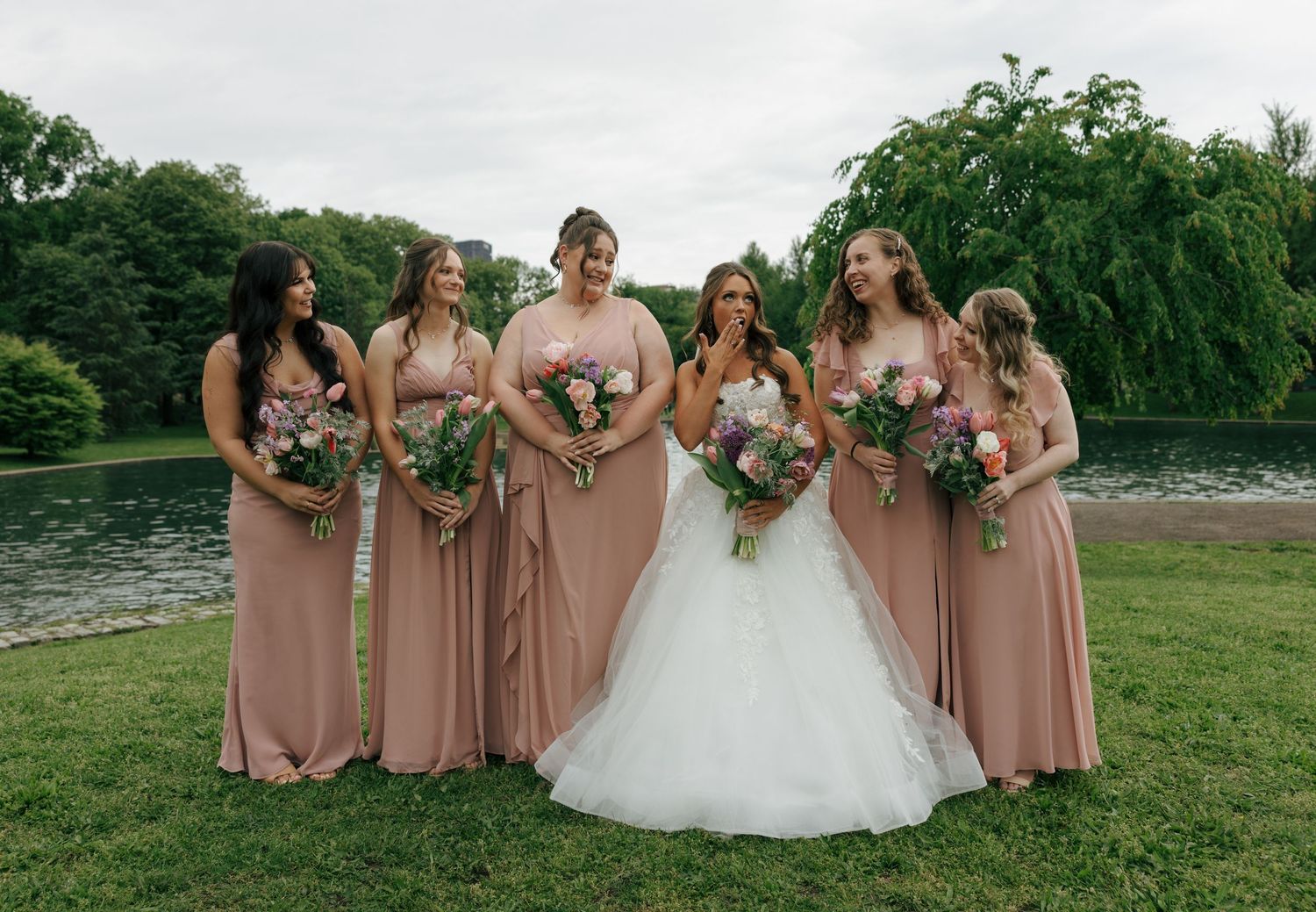 Bride in white gown with five bridesmaids in dusty rose dresses holding bouquets outdoors near a pond.