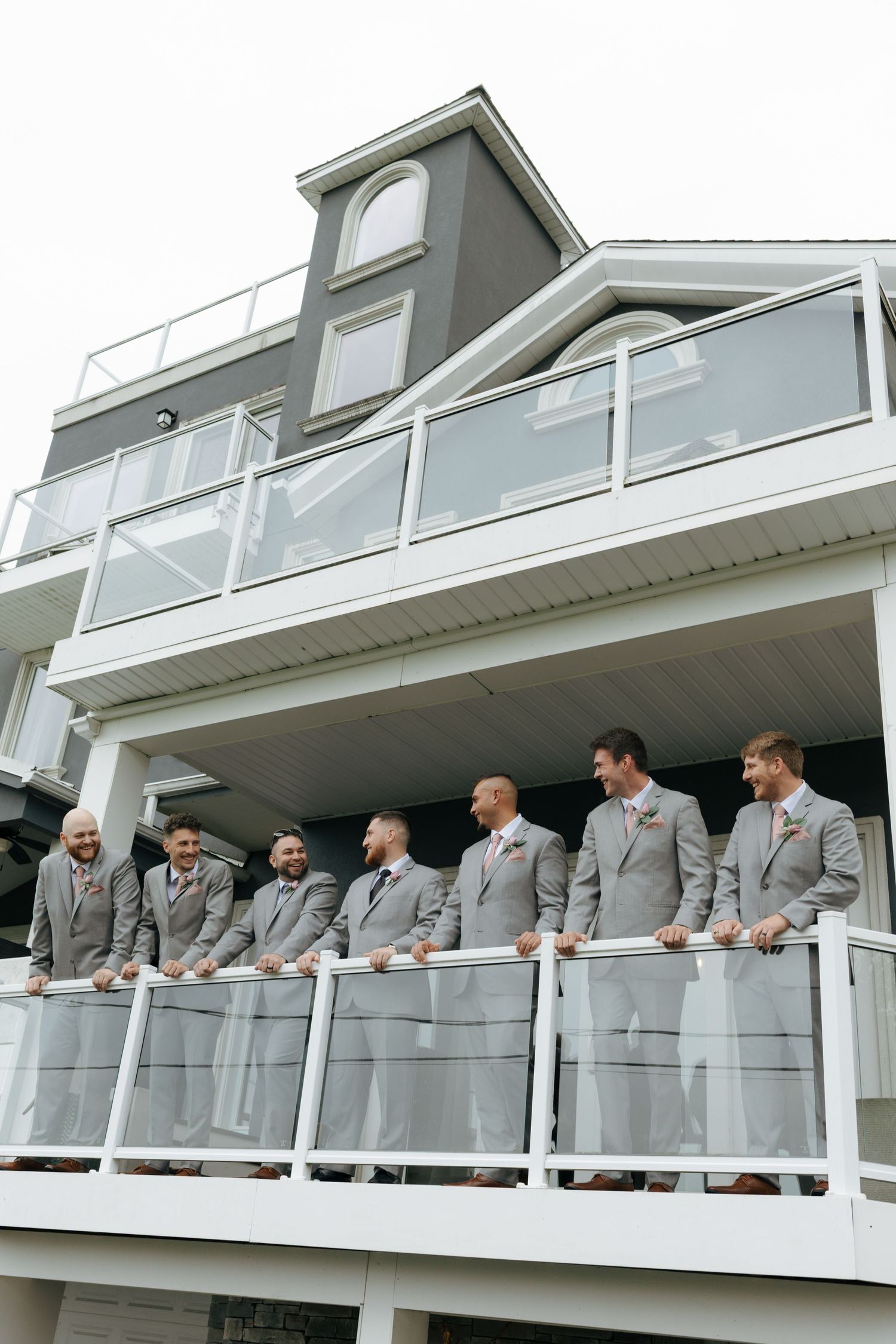 Wedding party groomsmen in matching gray suits pose on white balcony of elegant multi-story venue.