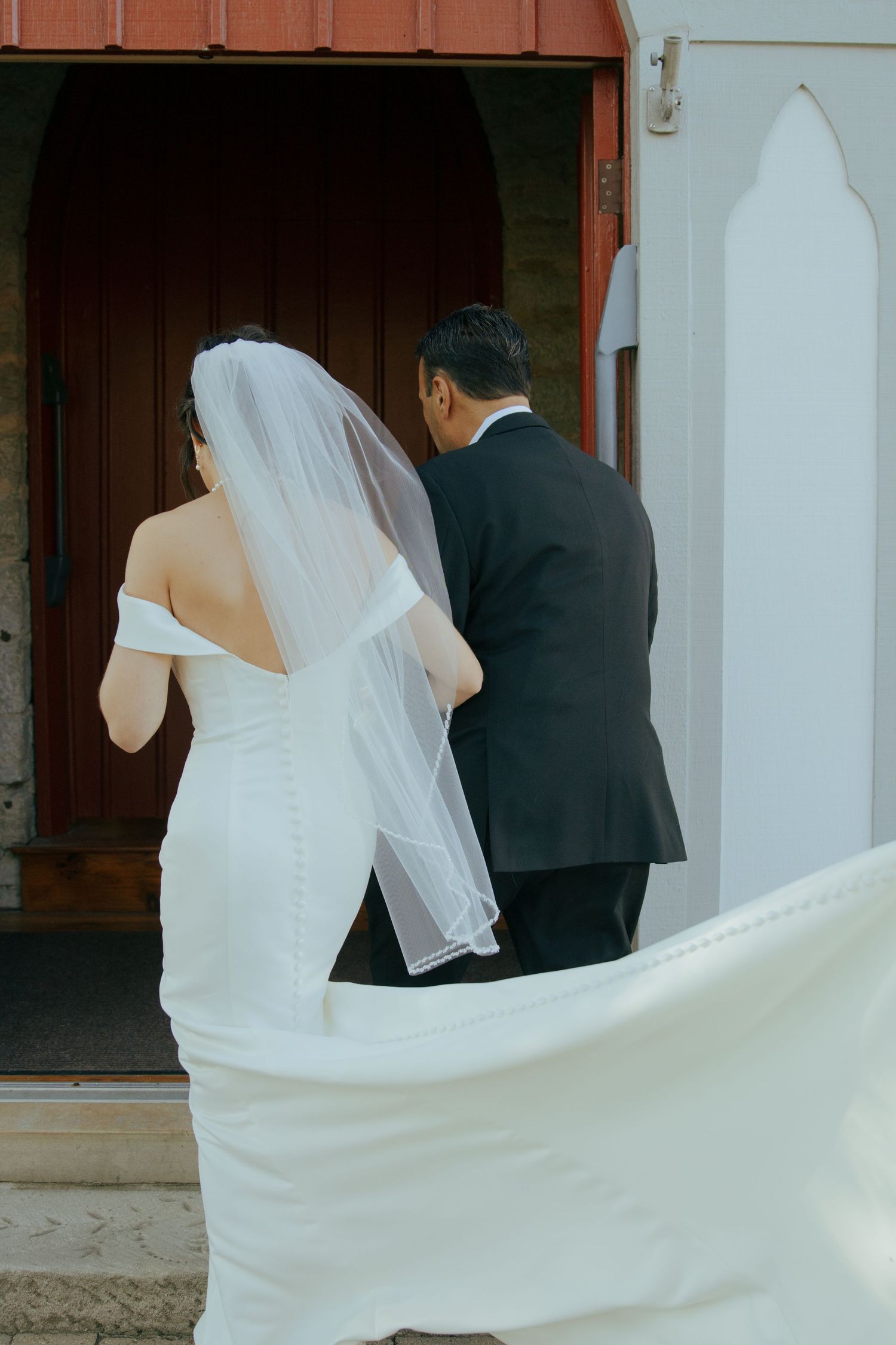 Bride in white off-shoulder gown with veil walking with groom in black suit toward church entrance.
