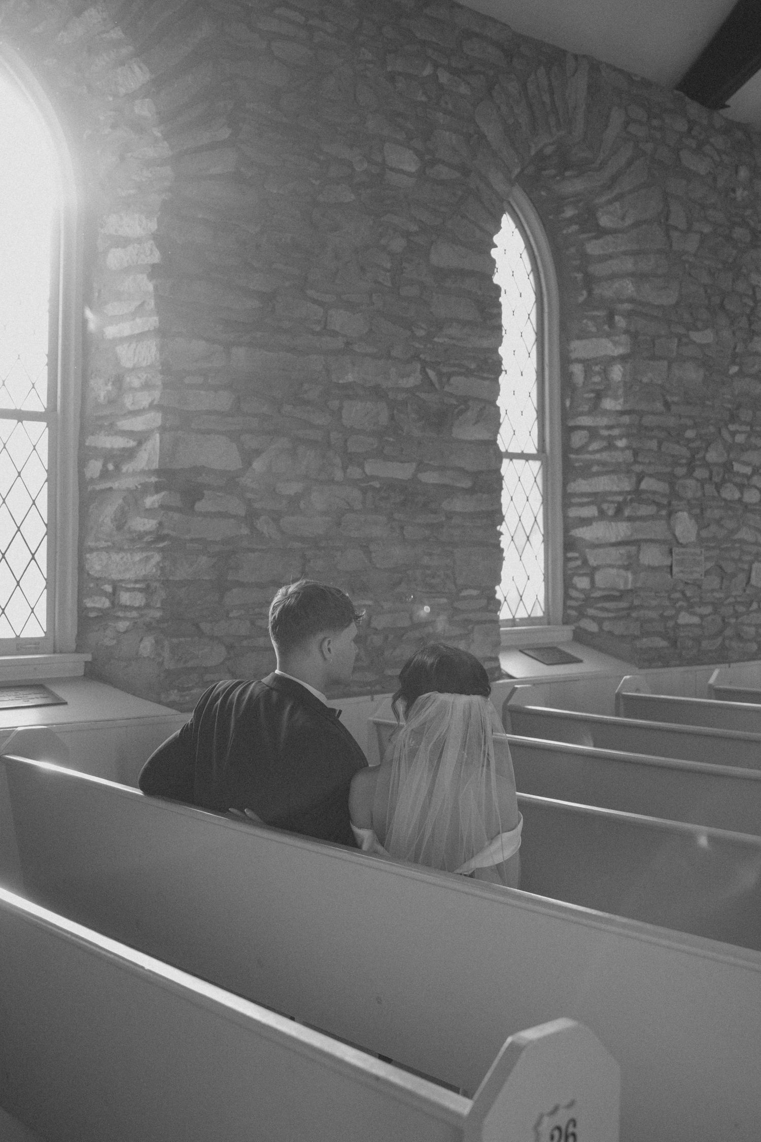 Black and white photo of couple sitting together in sunlit church pew during wedding.