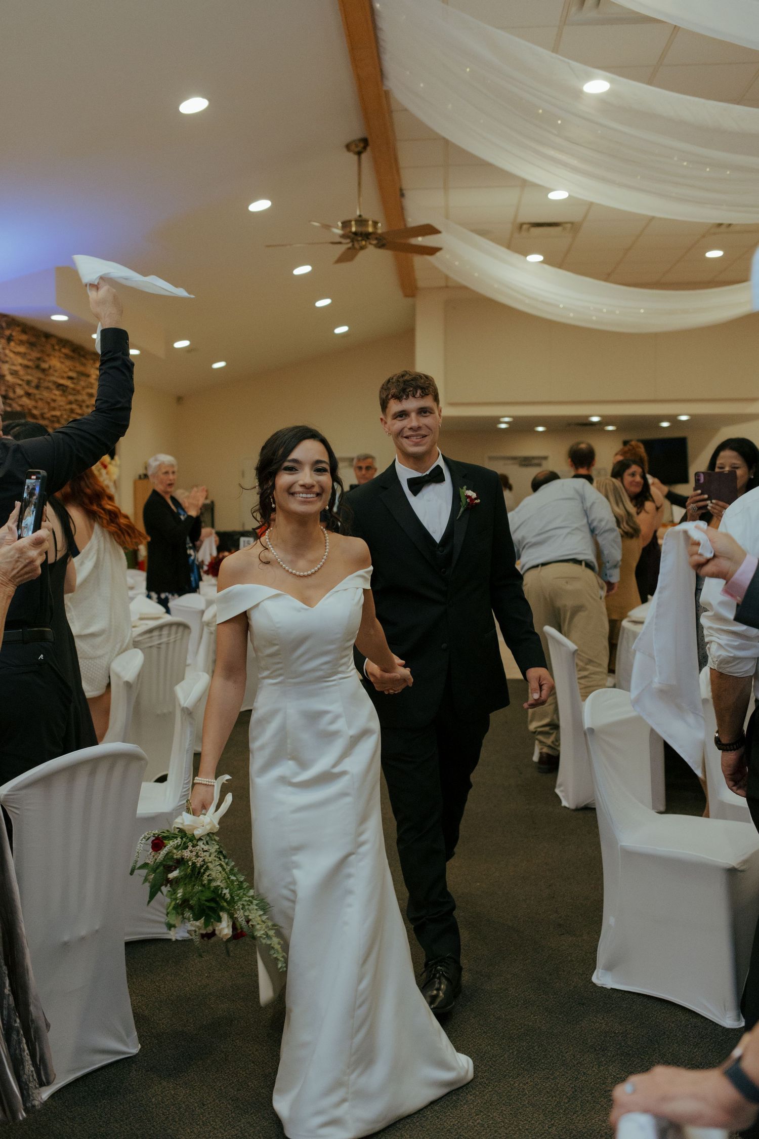 Newlyweds walk hand-in-hand down the aisle during their wedding reception entrance in a decorated banquet hall.