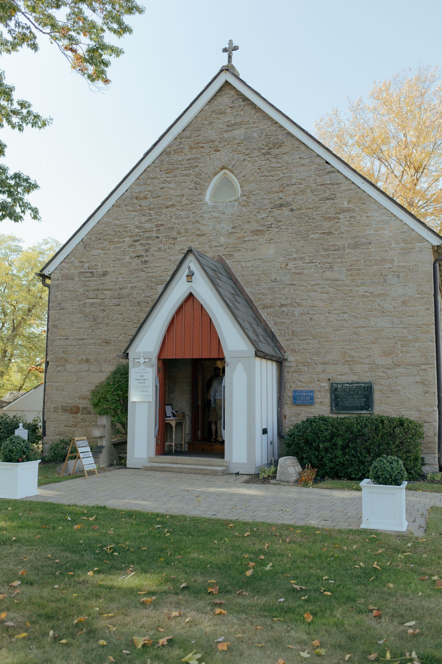 Historic stone church with white arched entrance, cross atop gabled roof, surrounded by autumn trees and manicured shrubs.