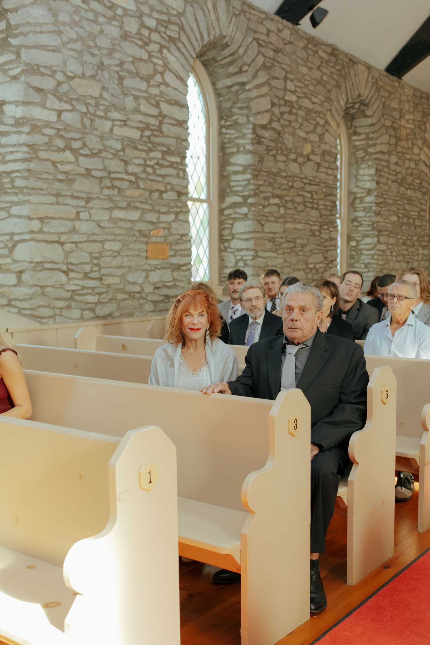 Guests seated in white wooden pews inside a historic stone church during a wedding ceremony.