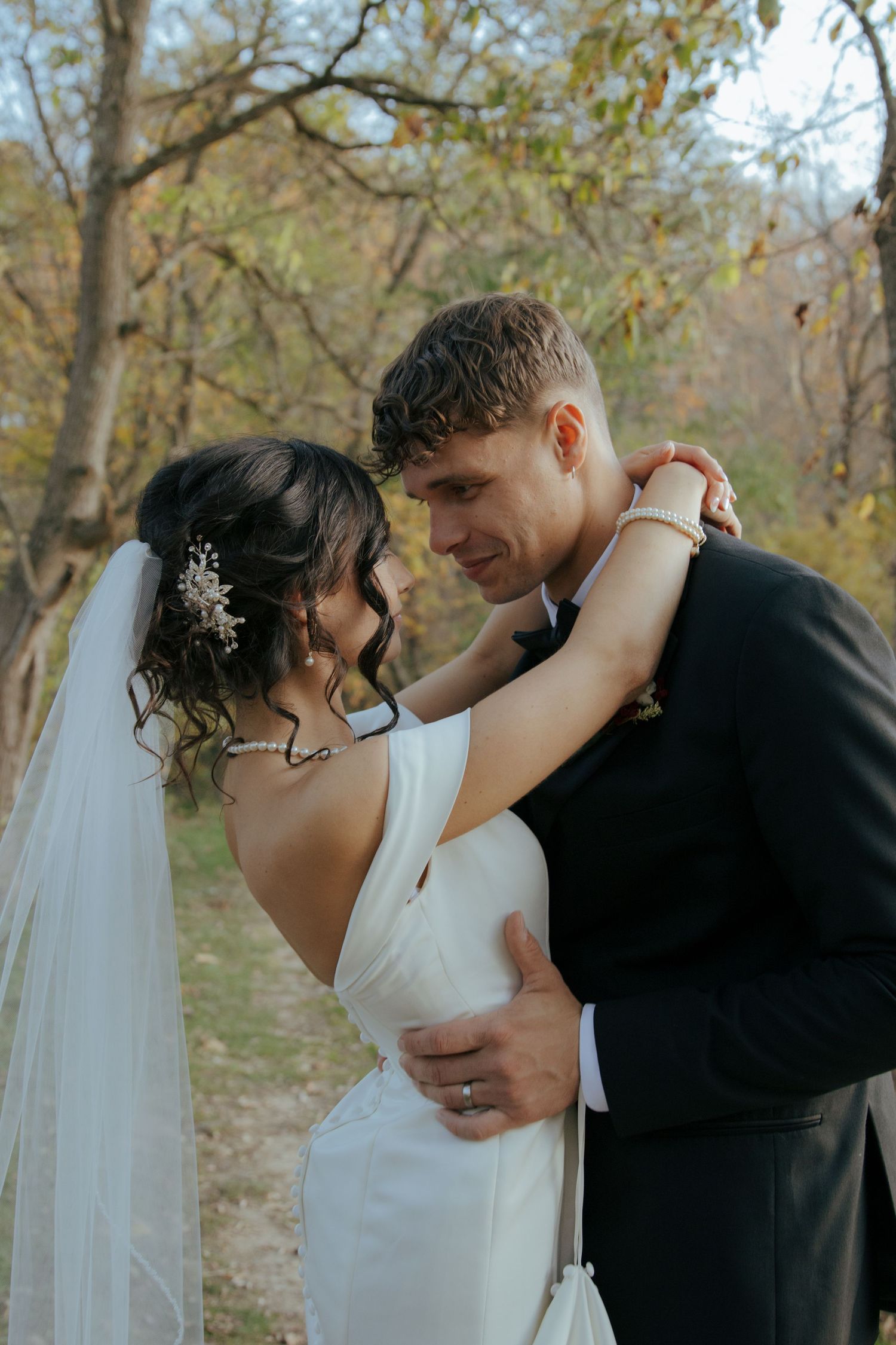 Bride in backless white gown and groom in black suit embrace tenderly among autumn trees outdoors.