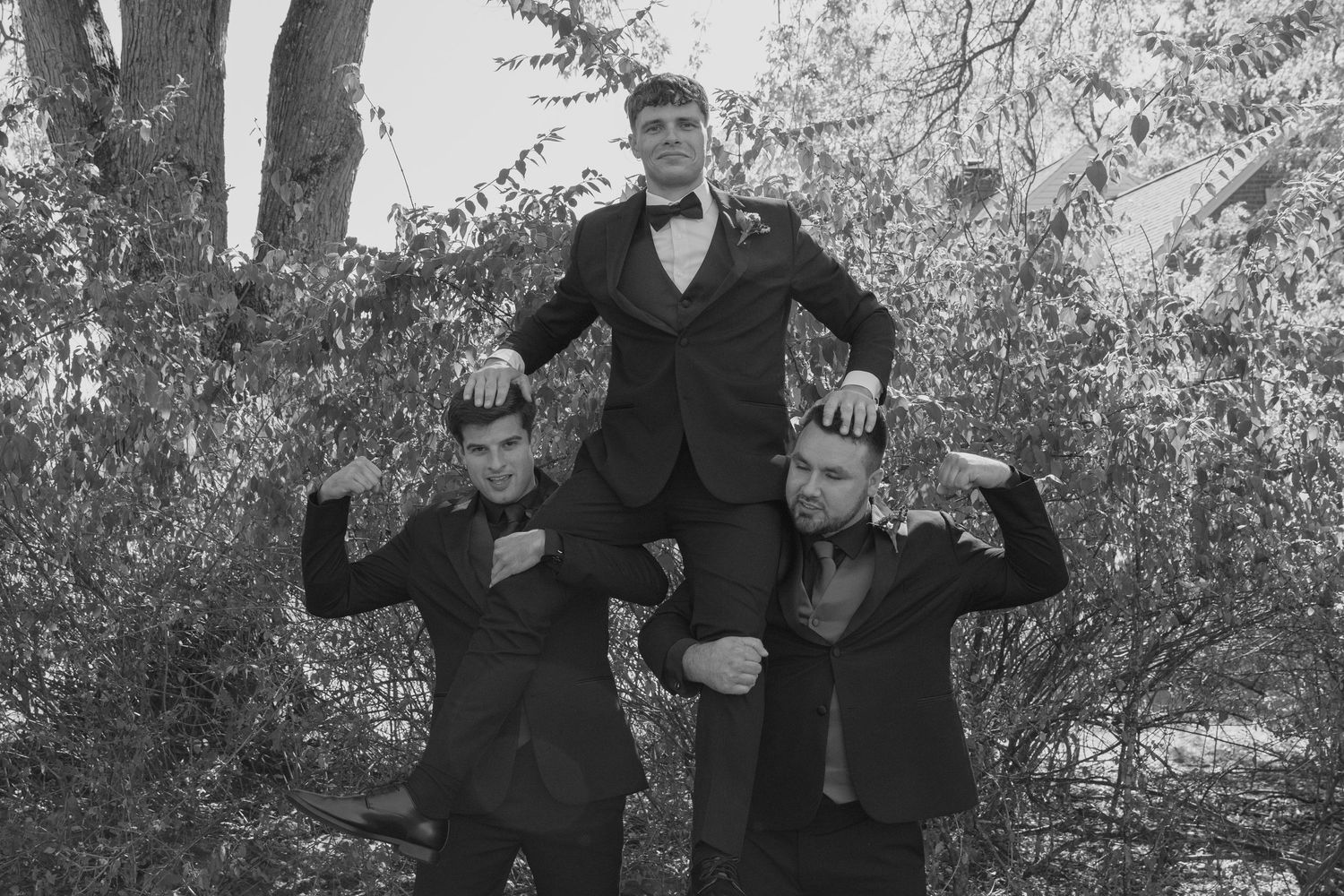 Three young men in black suits pose dramatically outdoors among trees in a black and white photo.
