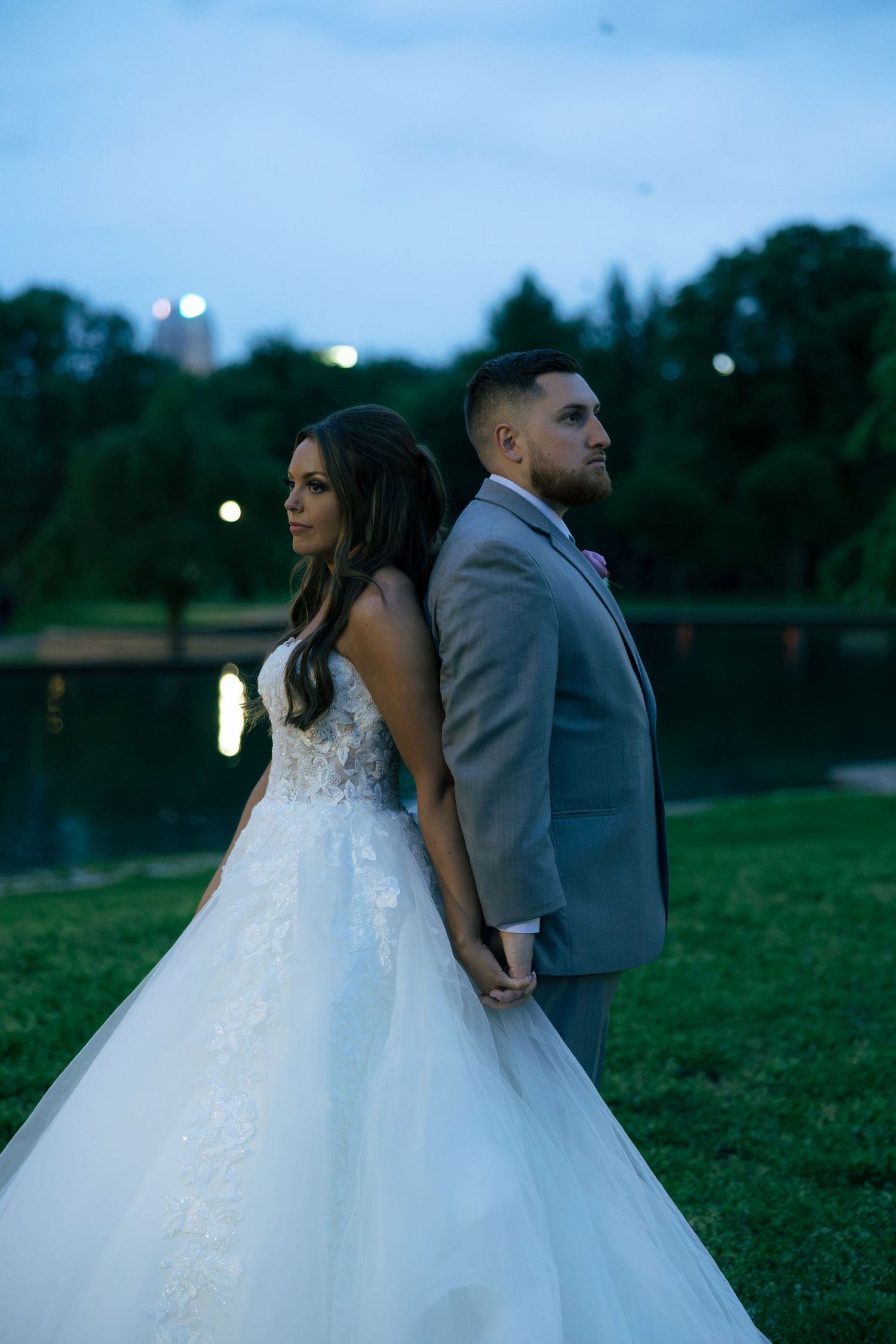 Bride and groom stand back to back holding hands by a pond at twilight in elegant wedding attire.