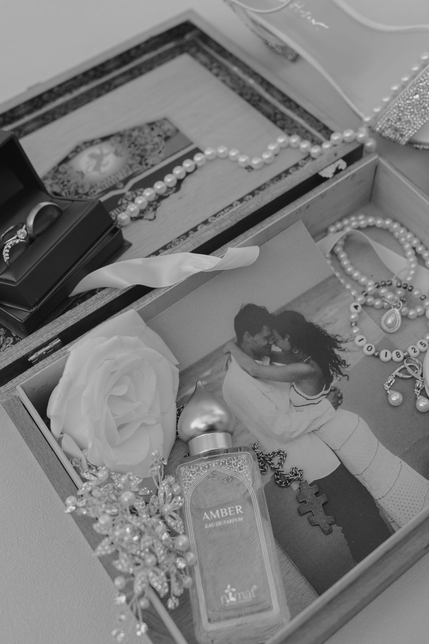 Black and white flat lay of bridal accessories in a box, including pearls, Amber perfume, and a floral decoration.