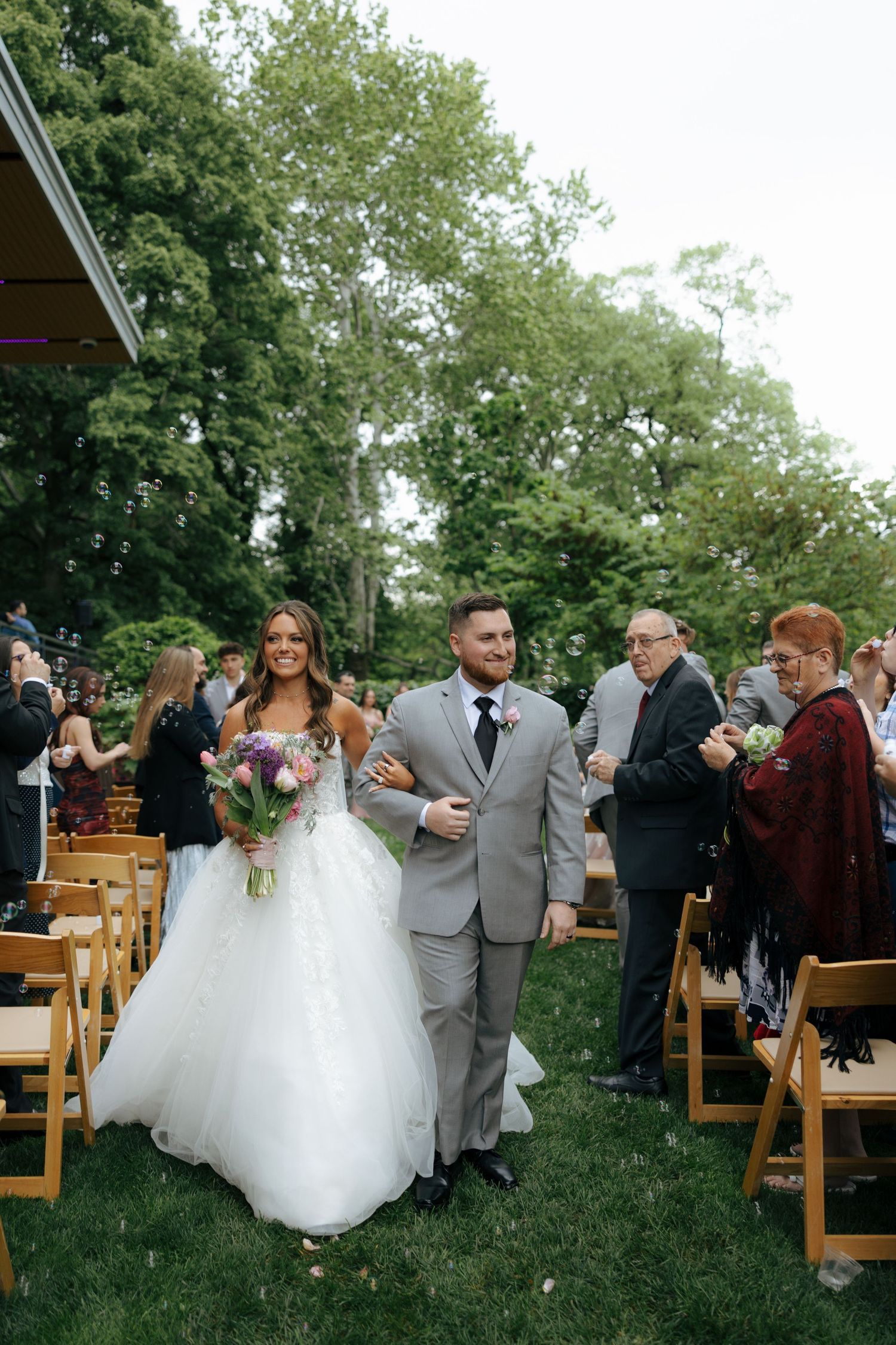 Newlyweds walk back down aisle after outdoor wedding ceremony, bride holding purple floral bouquet.