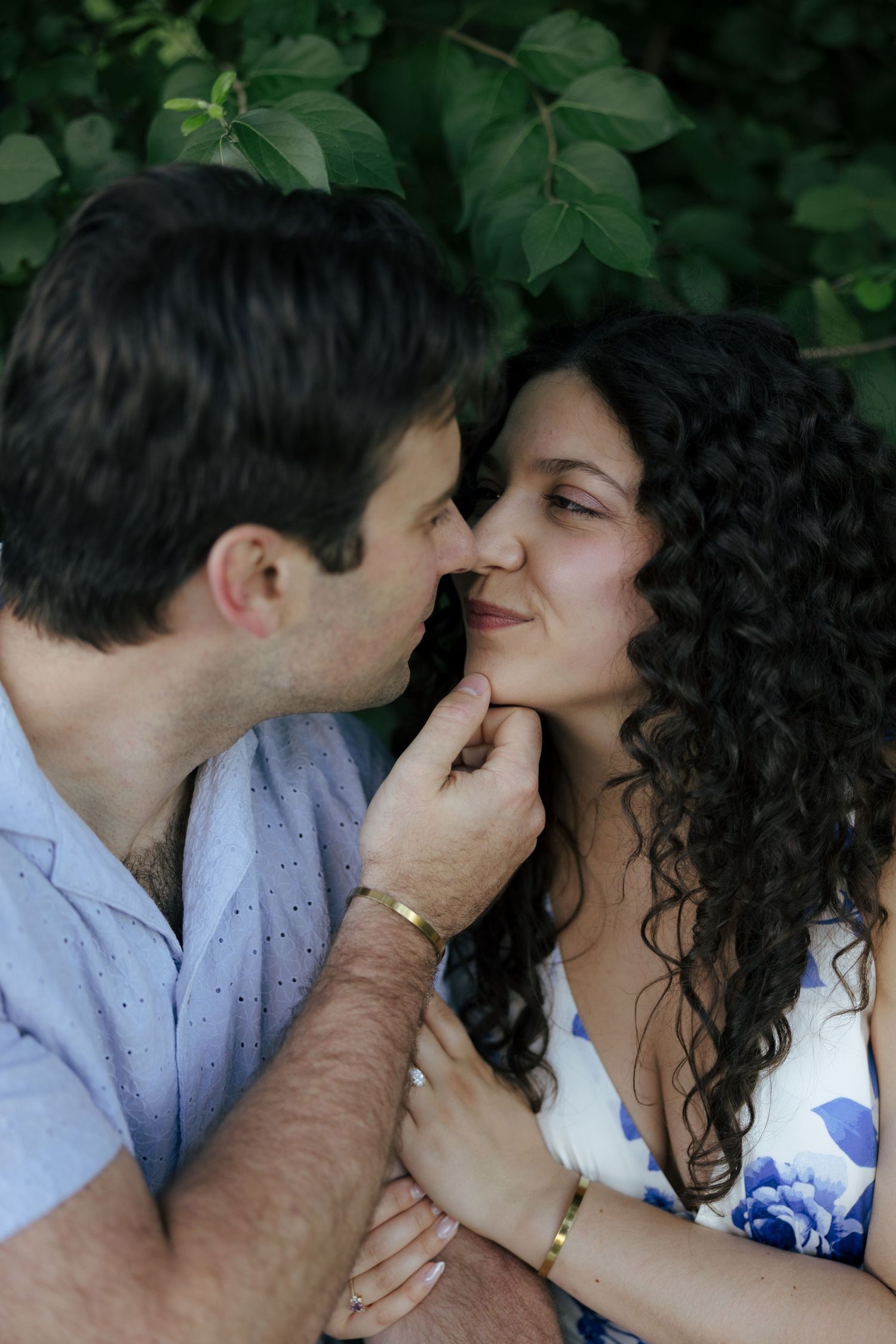 Man tenderly kissing woman's cheek while holding her face near lush green foliage.