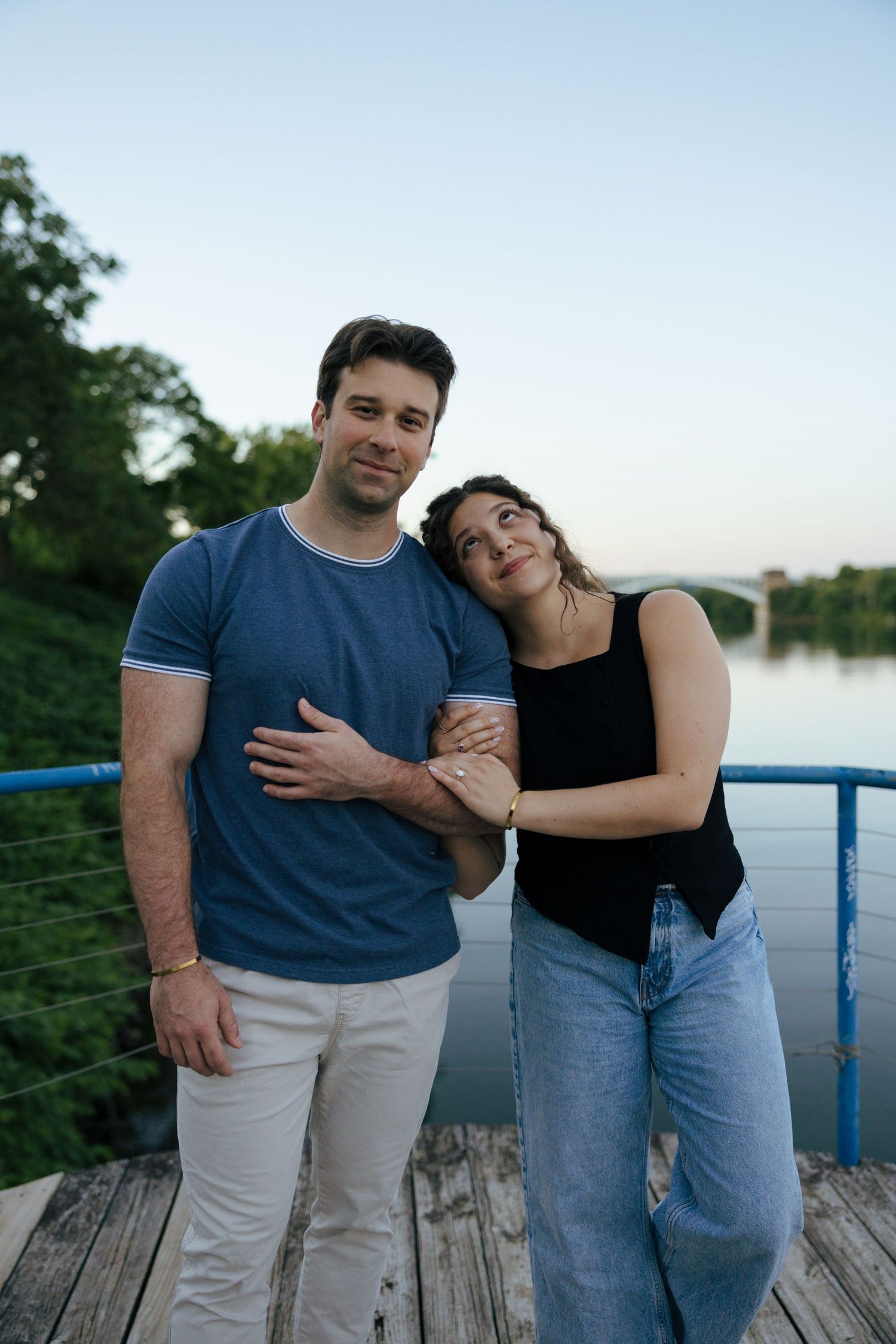A couple embraces on a wooden dock by a river at twilight, the woman leaning on the mans shoulder.