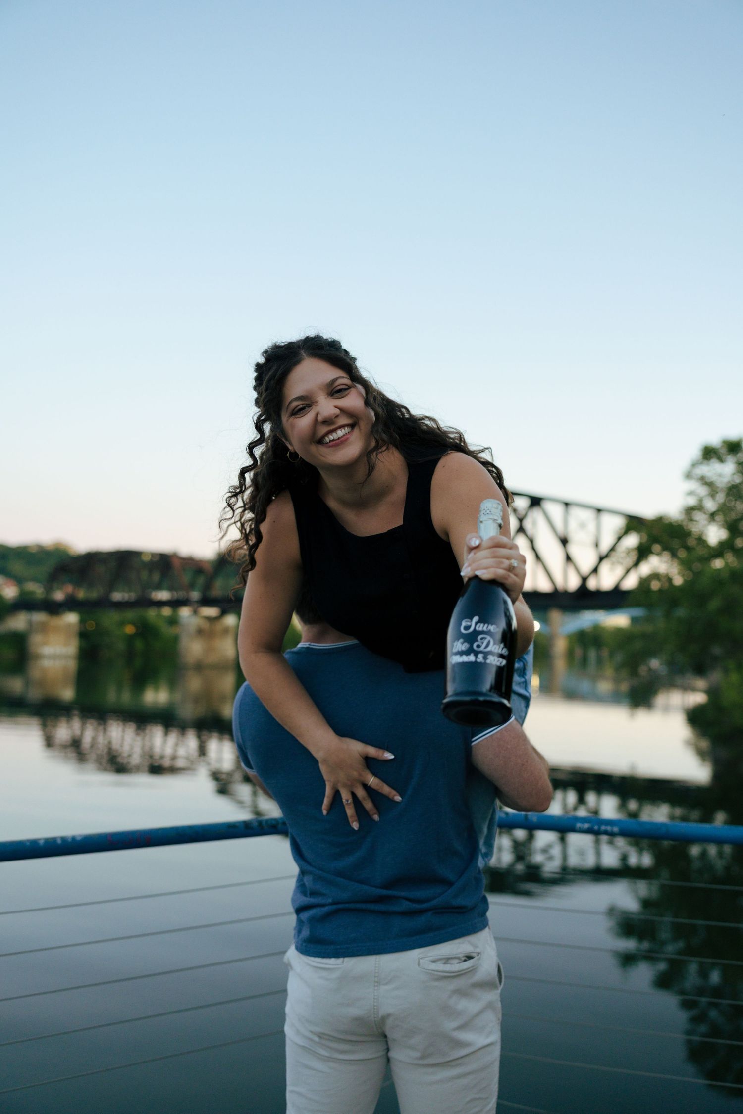 A joyful woman is lifted by her partner while holding a champagne bottle near a river bridge at sunset.