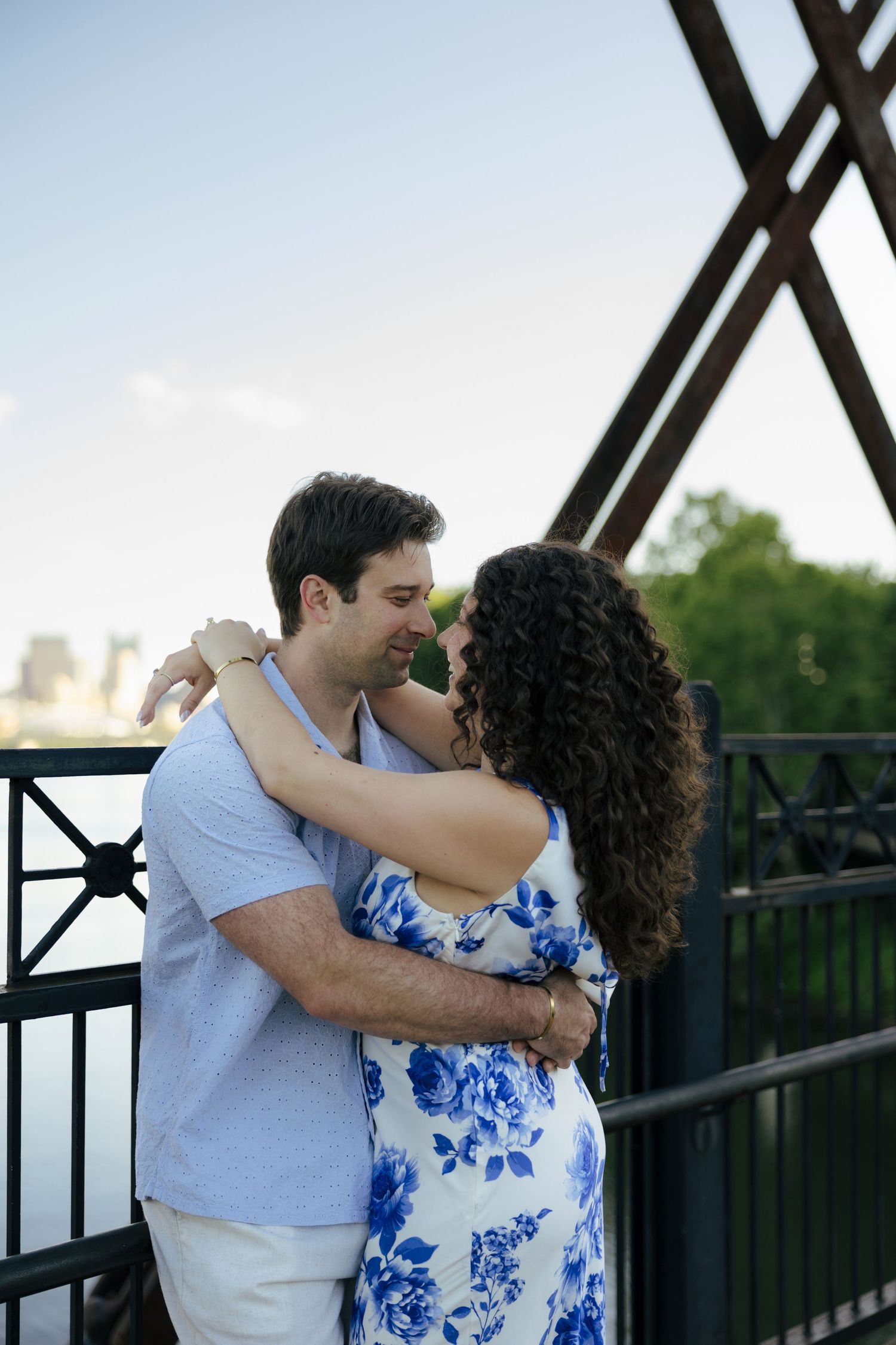 Romantic couple hugging face-to-face on a bridge, woman in blue floral dress, man in light blue shirt, greenery in background.