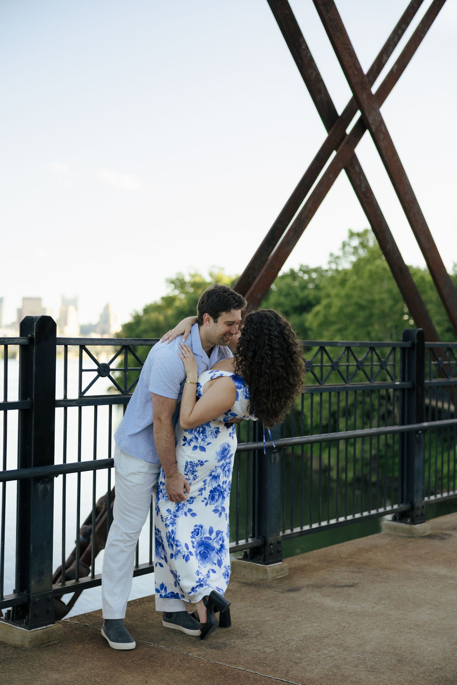 A couple shares a kiss on a bridge with rustic metal X-shaped structures in the background.