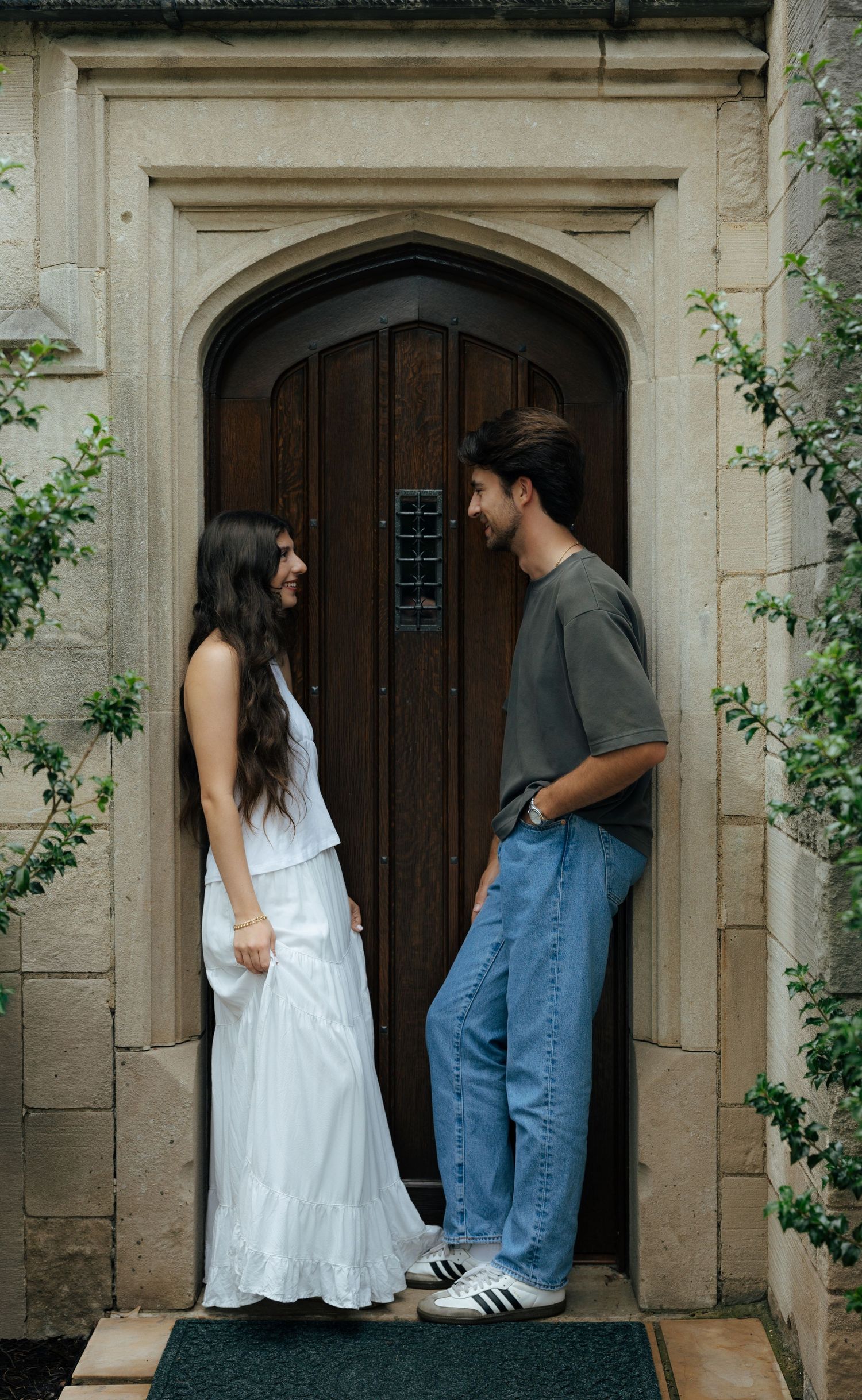 A couple smiles at each other while standing before an elegant arched wooden door on a stone building.