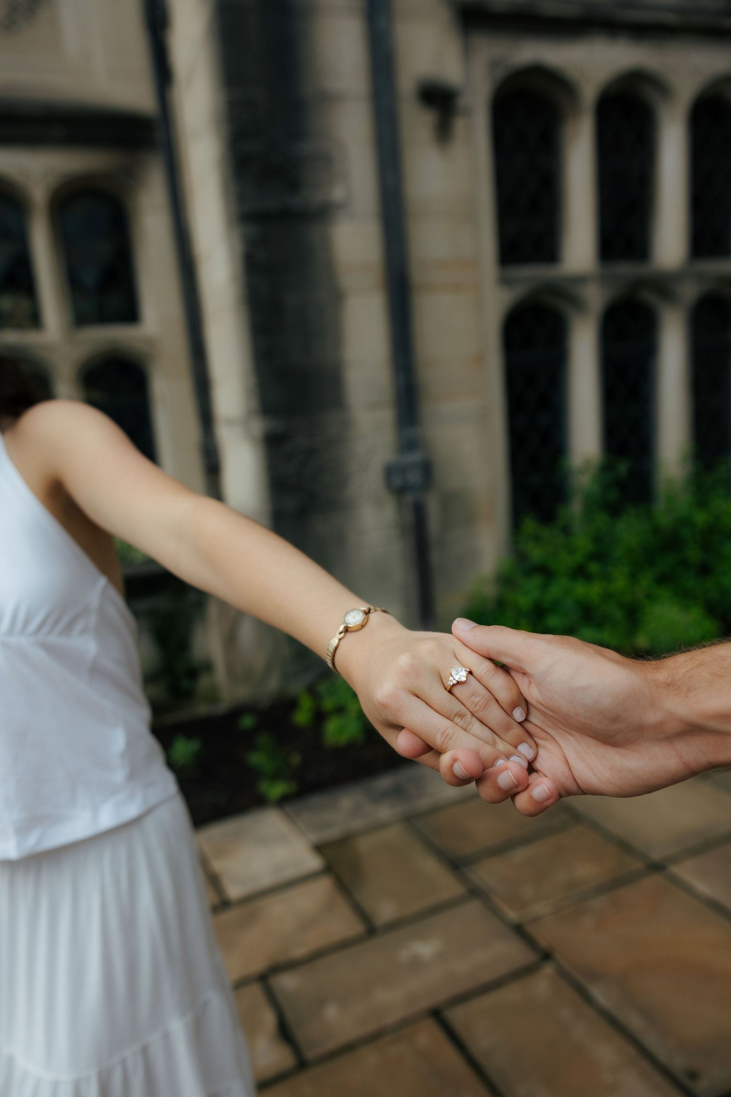 Close-up of engaged couple holding hands showing diamond engagement ring and gold bracelet outdoors.