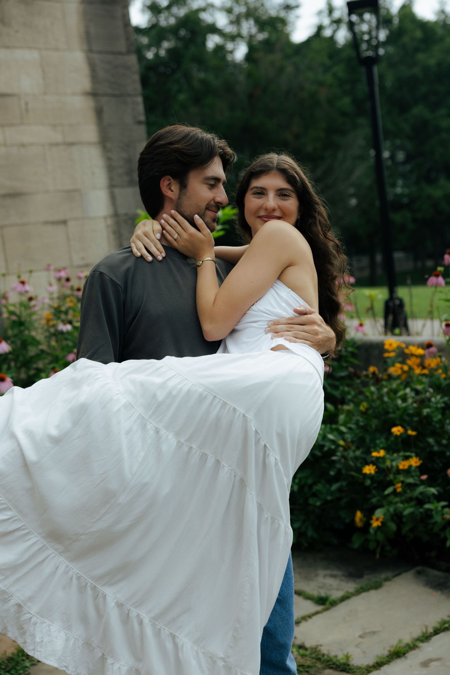 A man carries a woman in a white dress in his arms, both smiling, surrounded by colorful garden flowers.
