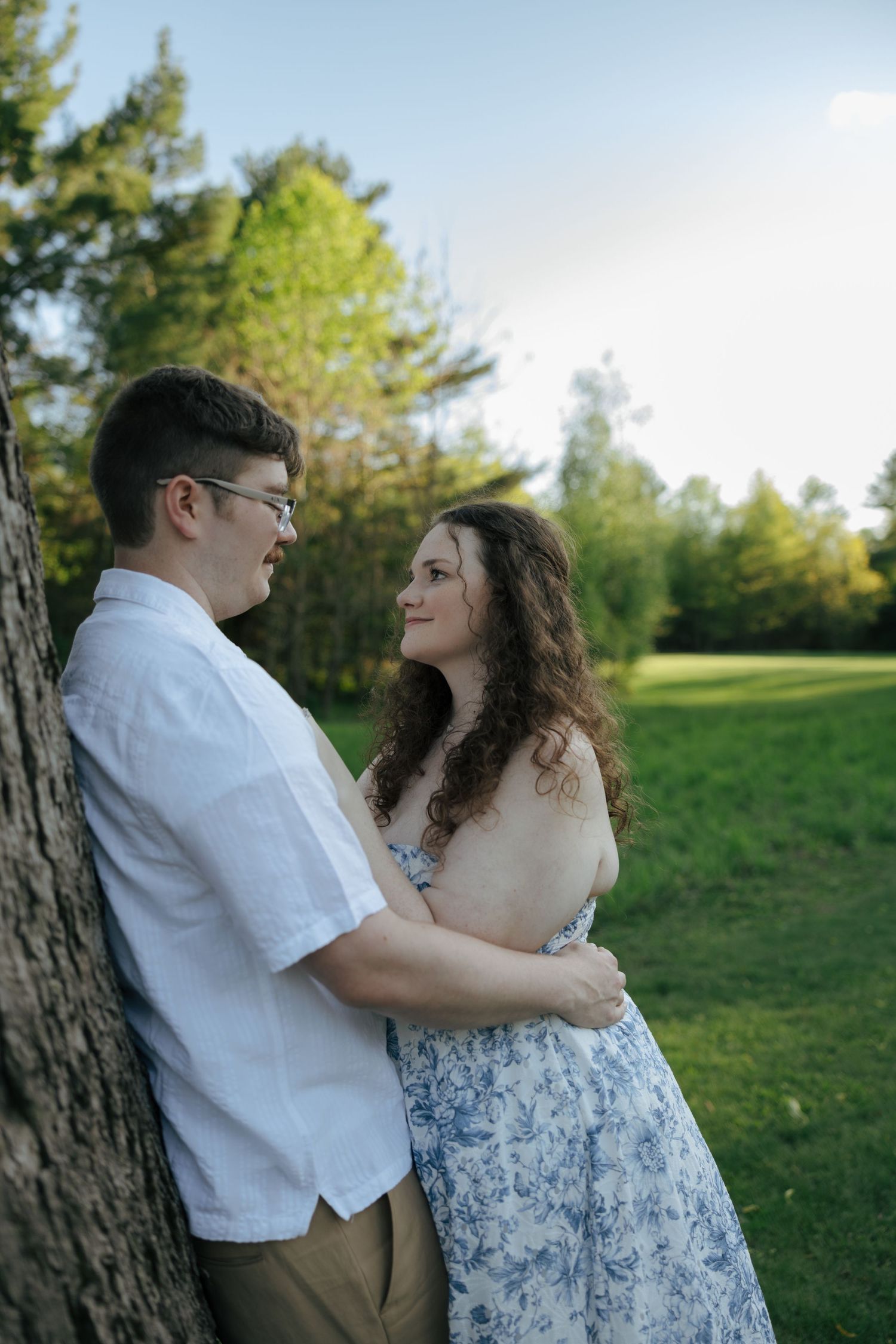 Couple embracing by a tree in a sunny green park, man in white shirt and woman in floral dress gazing at each other.