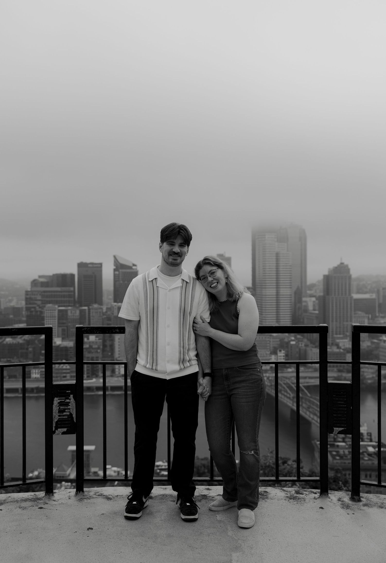 Black and white photo of a couple posing on a rooftop overlook with a city skyline in the background.