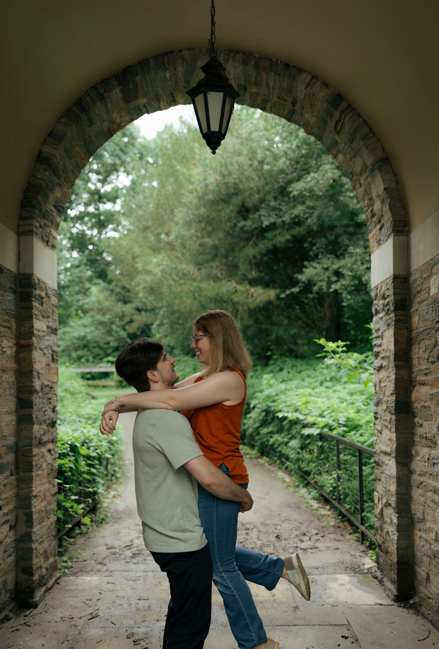 Couple embracing and laughing under a stone archway surrounded by lush green trees.