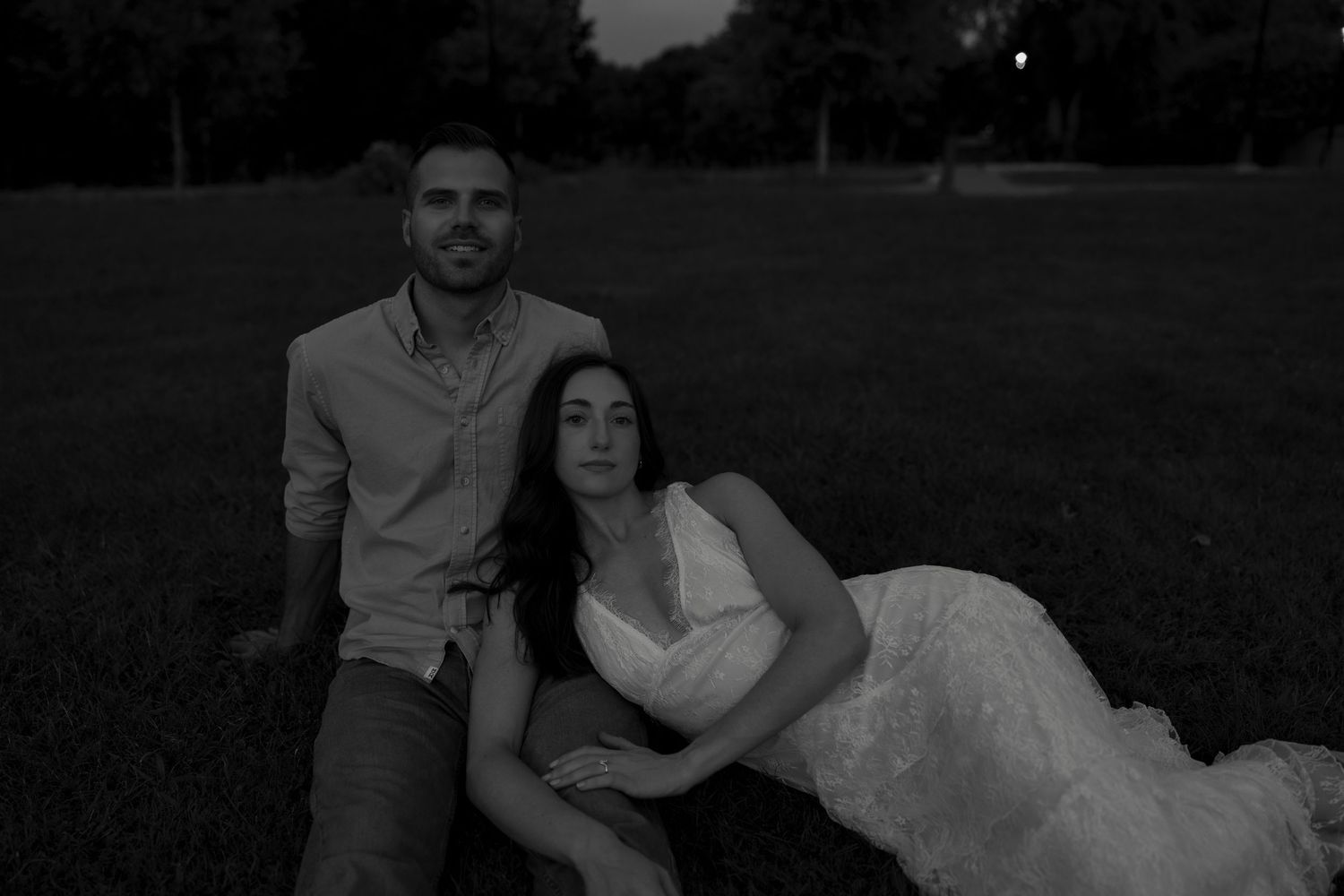 Black and white photo of engaged couple sitting together on lawn during twilight session.