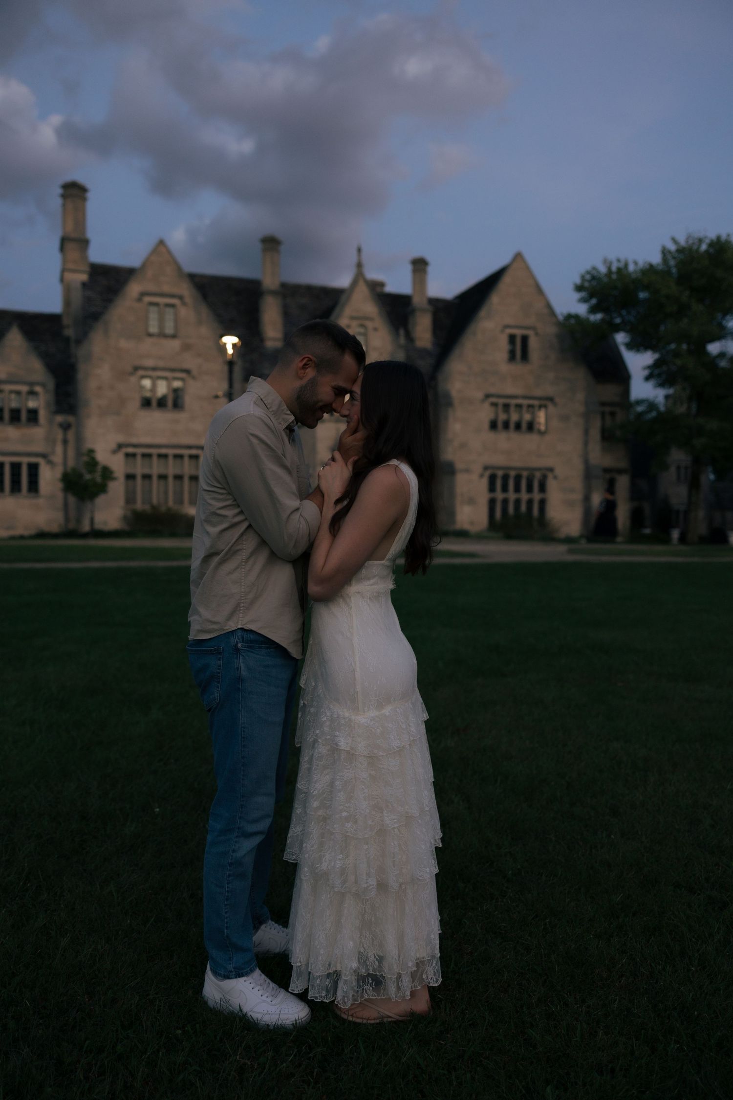 Couple embracing at dusk in front of a grand stone manor house on a lush green lawn.