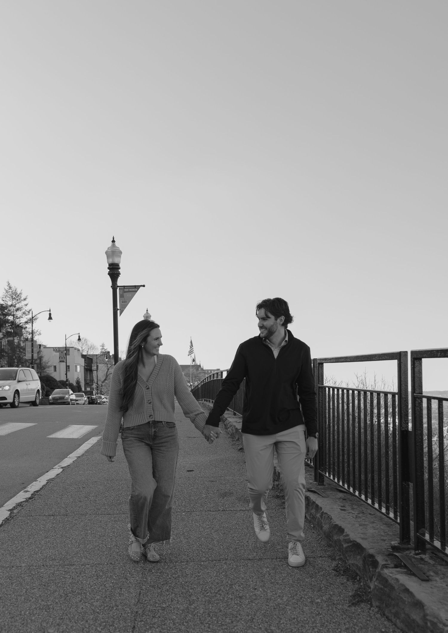 Black and white photo of a couple walking hand in hand along a waterfront path with street lamps.