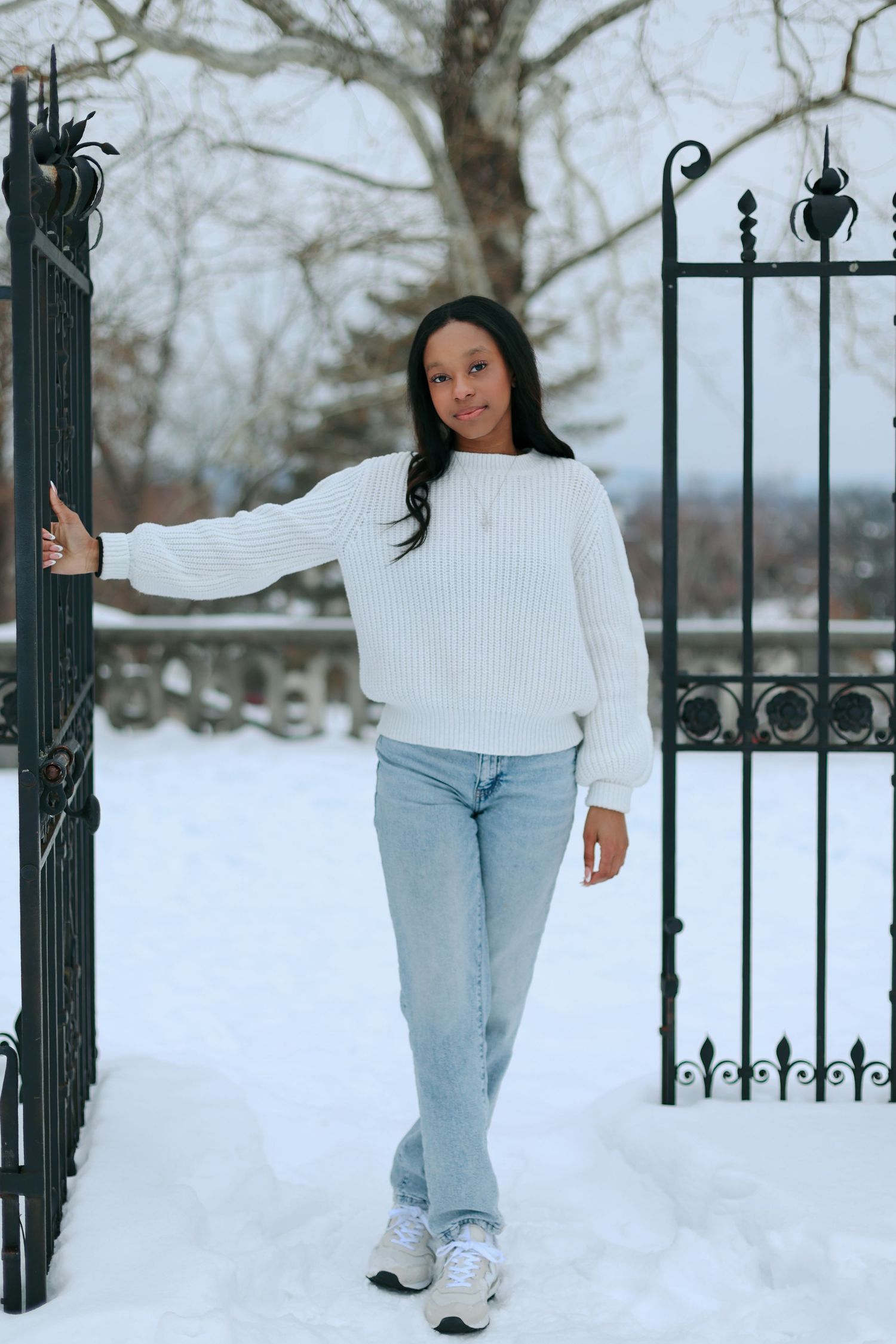 A person in a white sweater and jeans poses in front of a snow-covered wrought iron gate during winter.