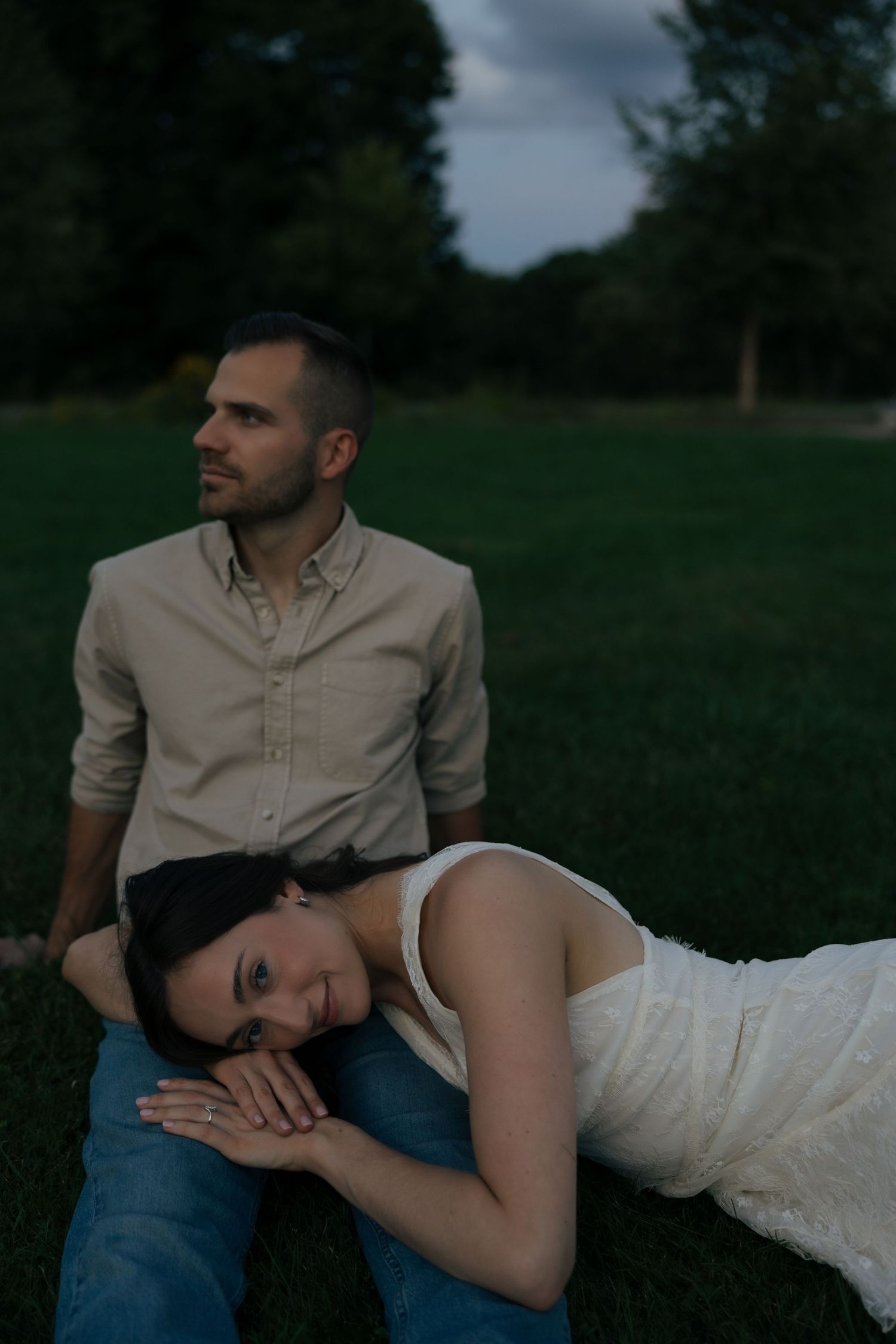 A woman in a white dress rests her head on a mans lap as they sit on green grass at dusk.