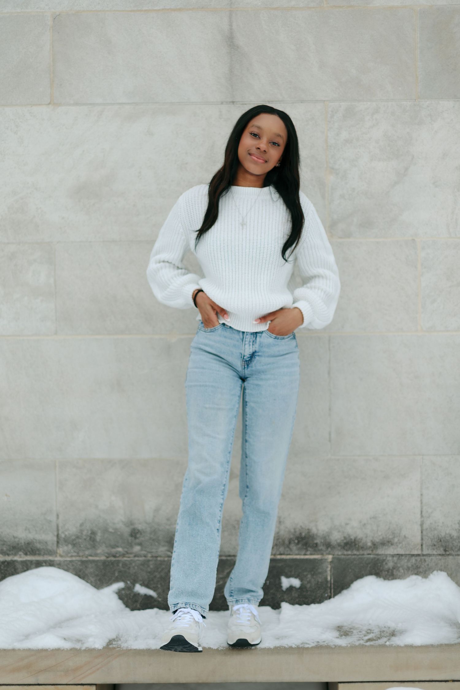 Young woman in white knit sweater and light blue jeans standing against a stone wall with snow.