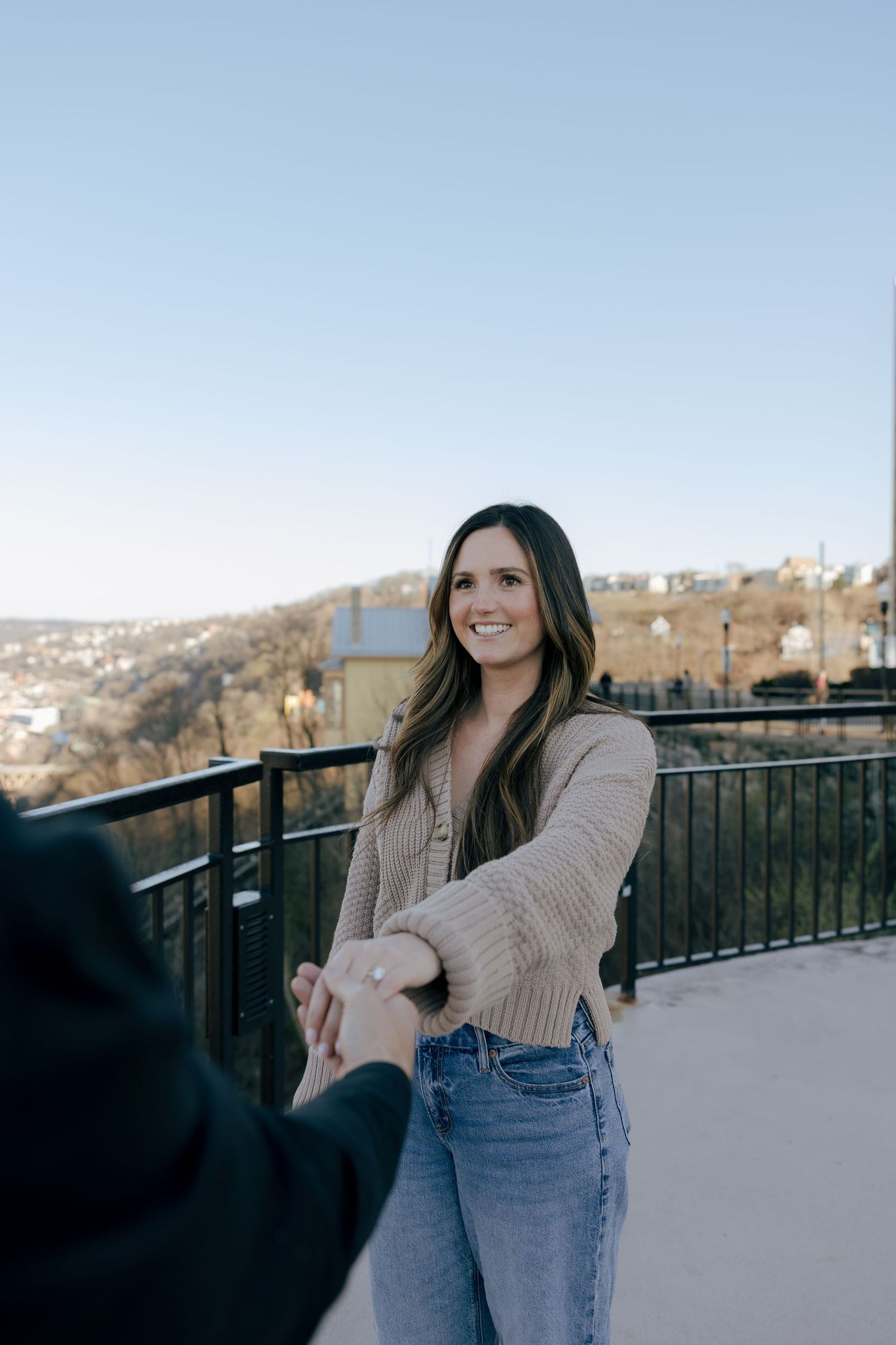 Couple holding hands on an outdoor observation deck overlooking a scenic city view on a sunny day.