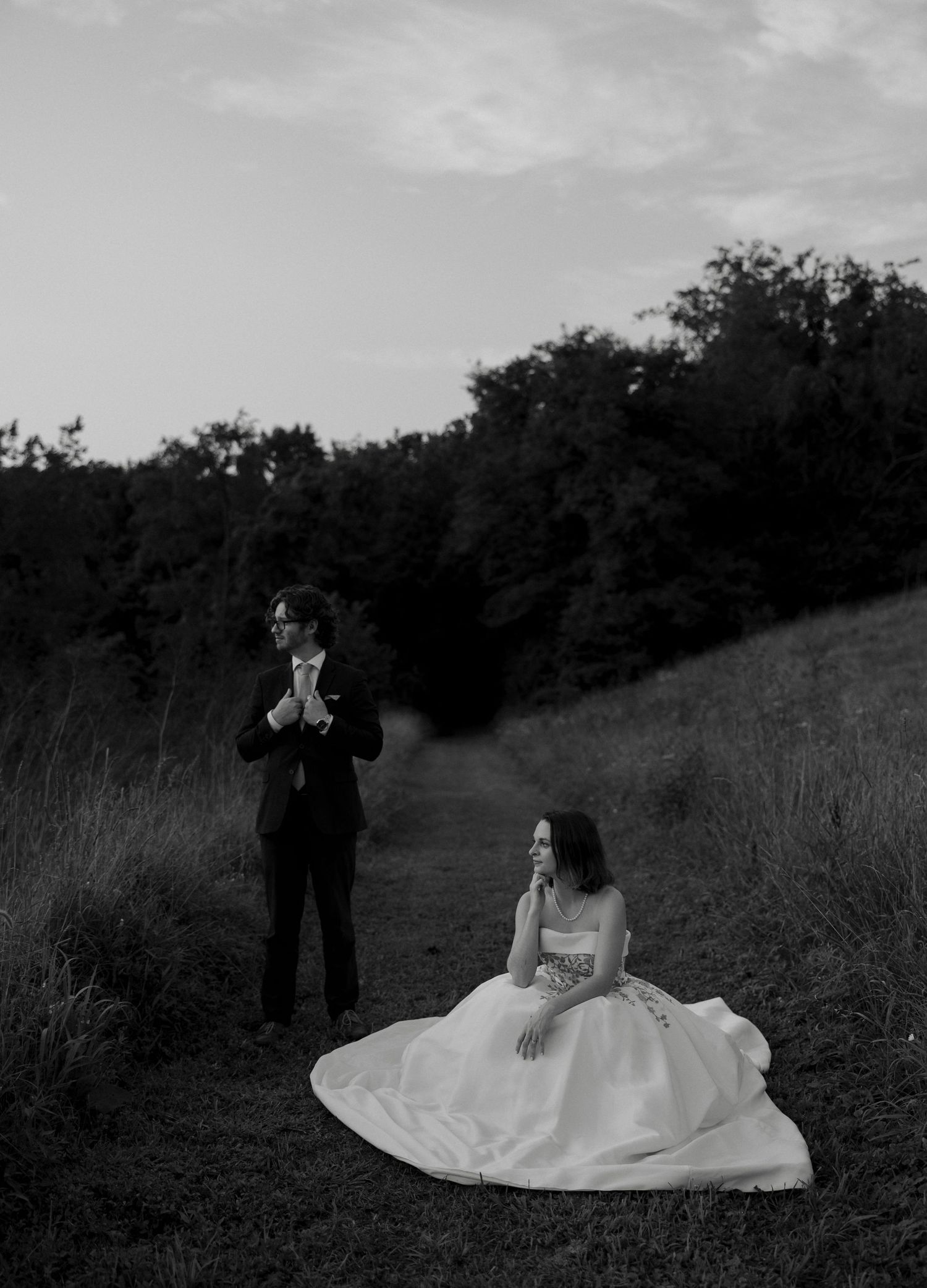 Black and white photo of bride seated on hillside path and groom standing in field at dusk.