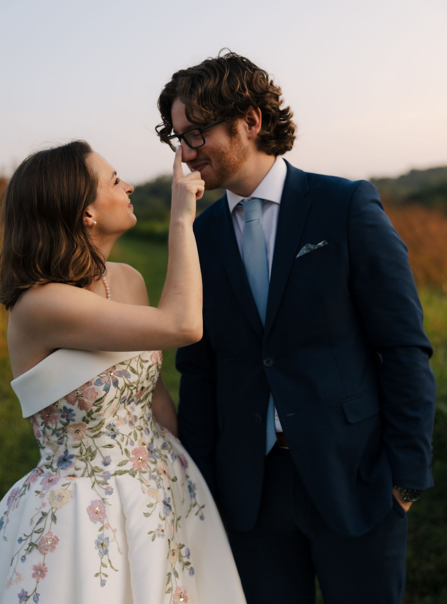Bride in floral wedding gown playfully touches grooms glasses as they smile at each other outdoors.