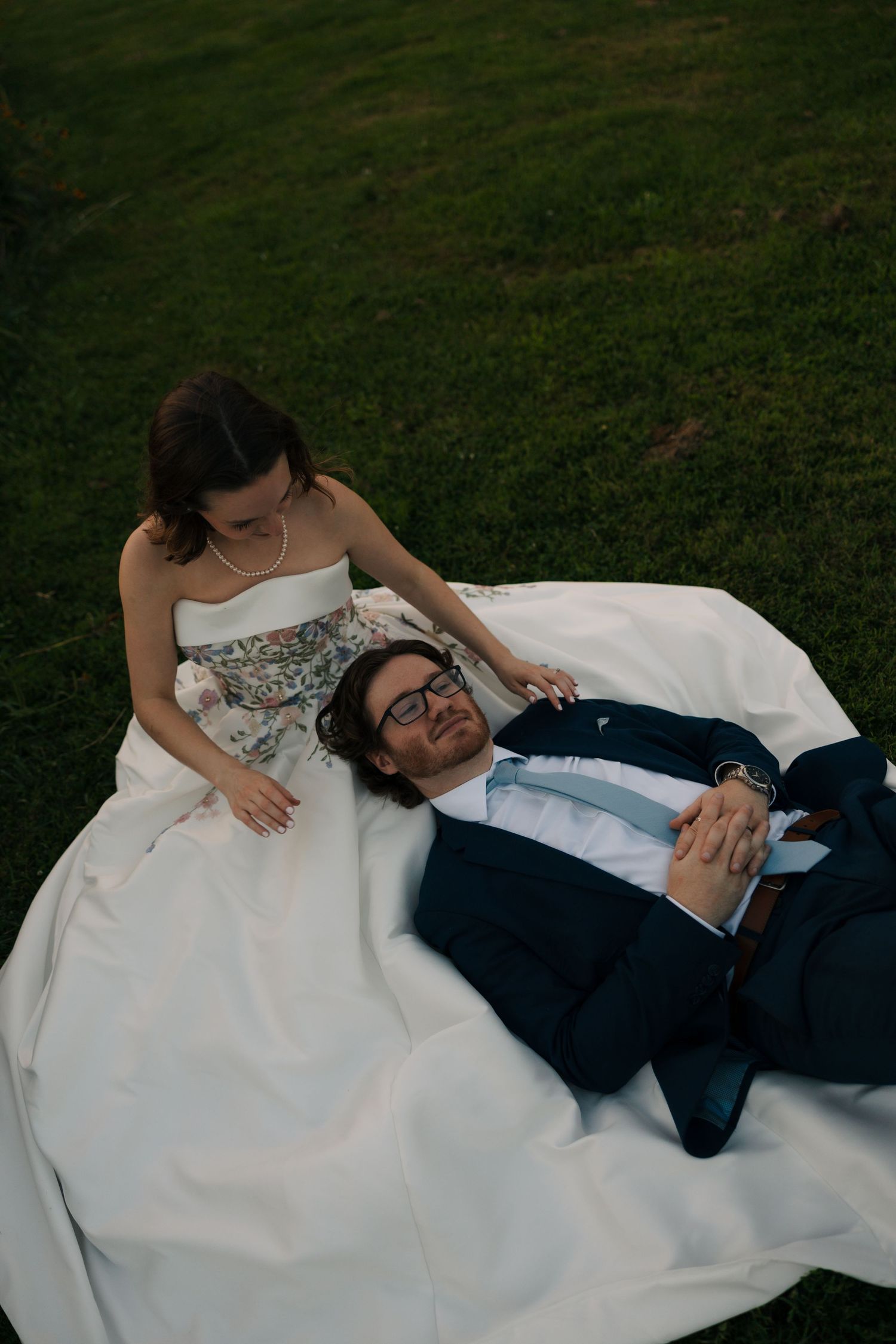 Bride and groom lying on grass, bride in floral corset gown holding hands at dusk.