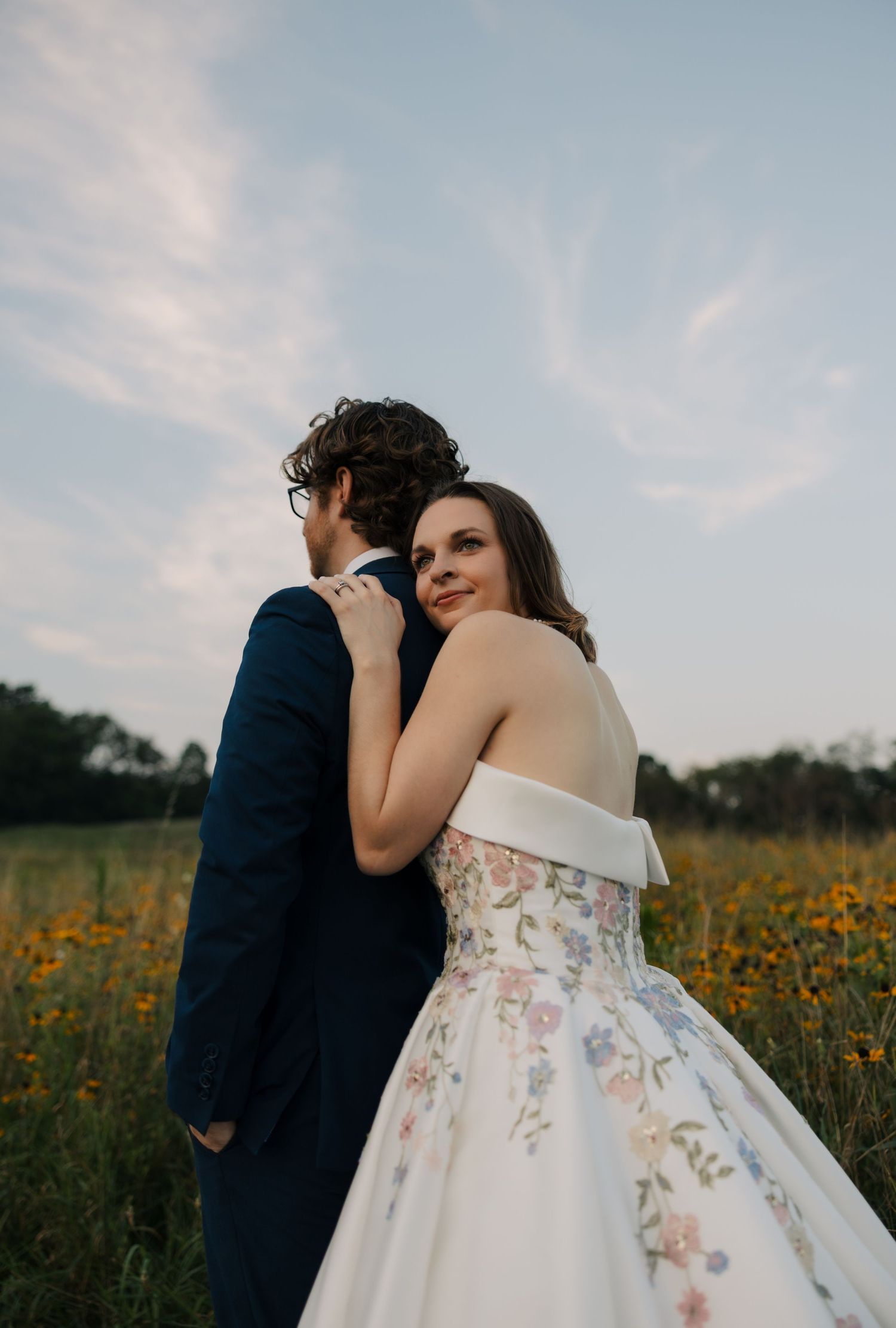 Bride in white floral ball gown embraces groom in navy suit in a sunflower field at sunset.