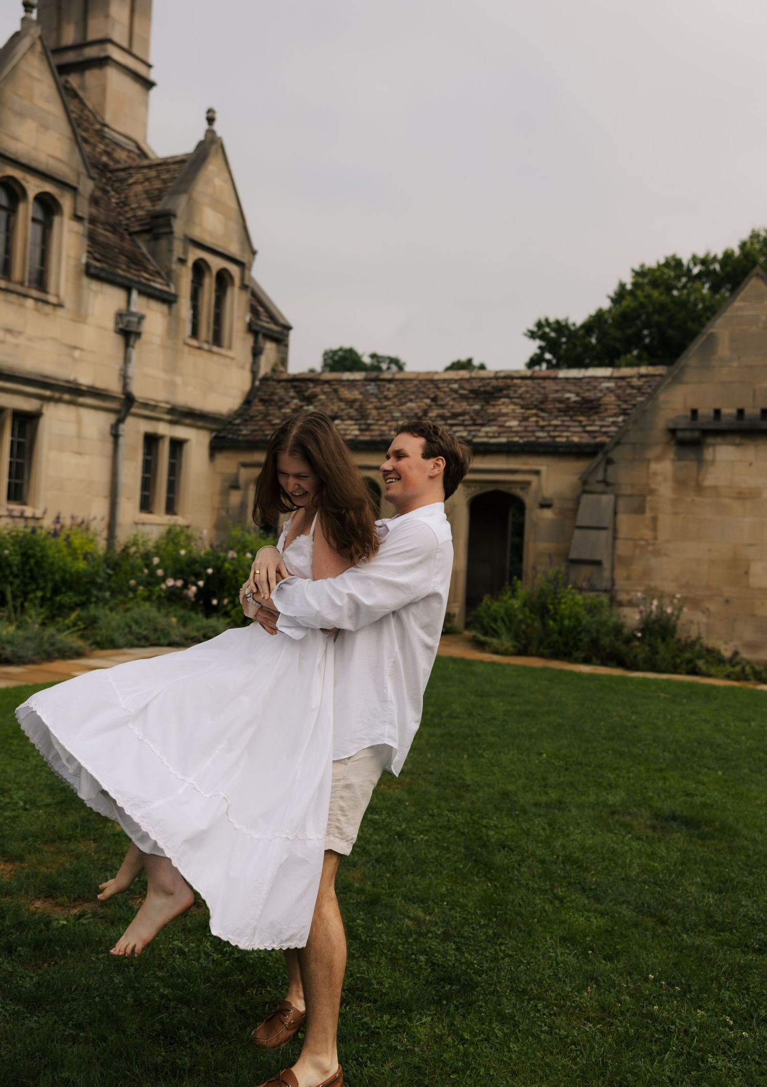 Joyful man lifting woman in white dress while spinning on green lawn in front of a historic manor.