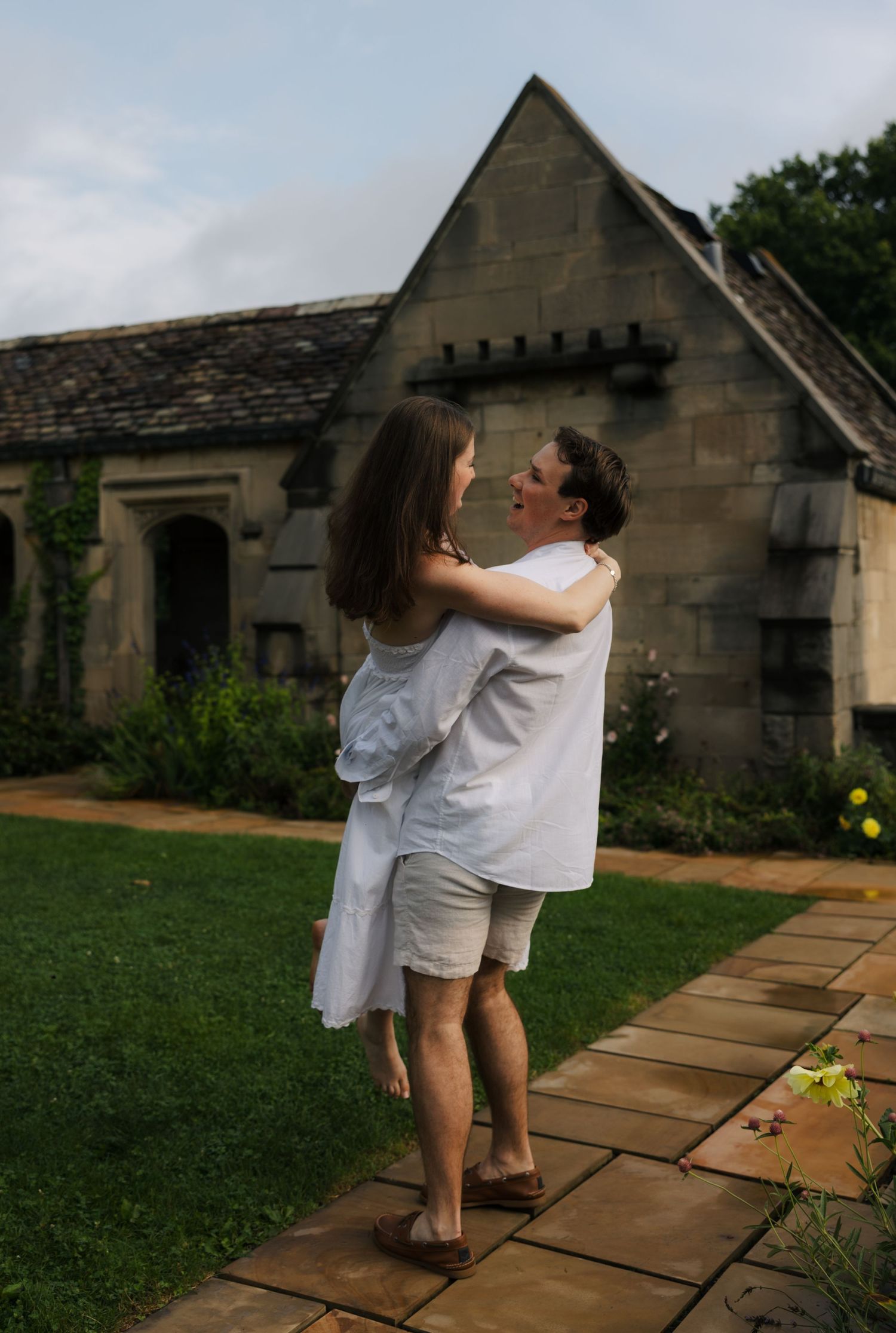Couple in white outfits embracing on garden path before stone cottage.
