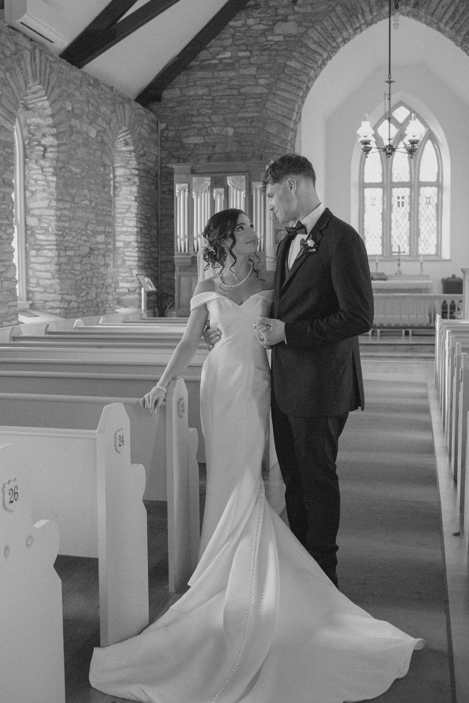 Black and white photo of bride in mermaid gown and groom standing together inside a rustic stone chapel.