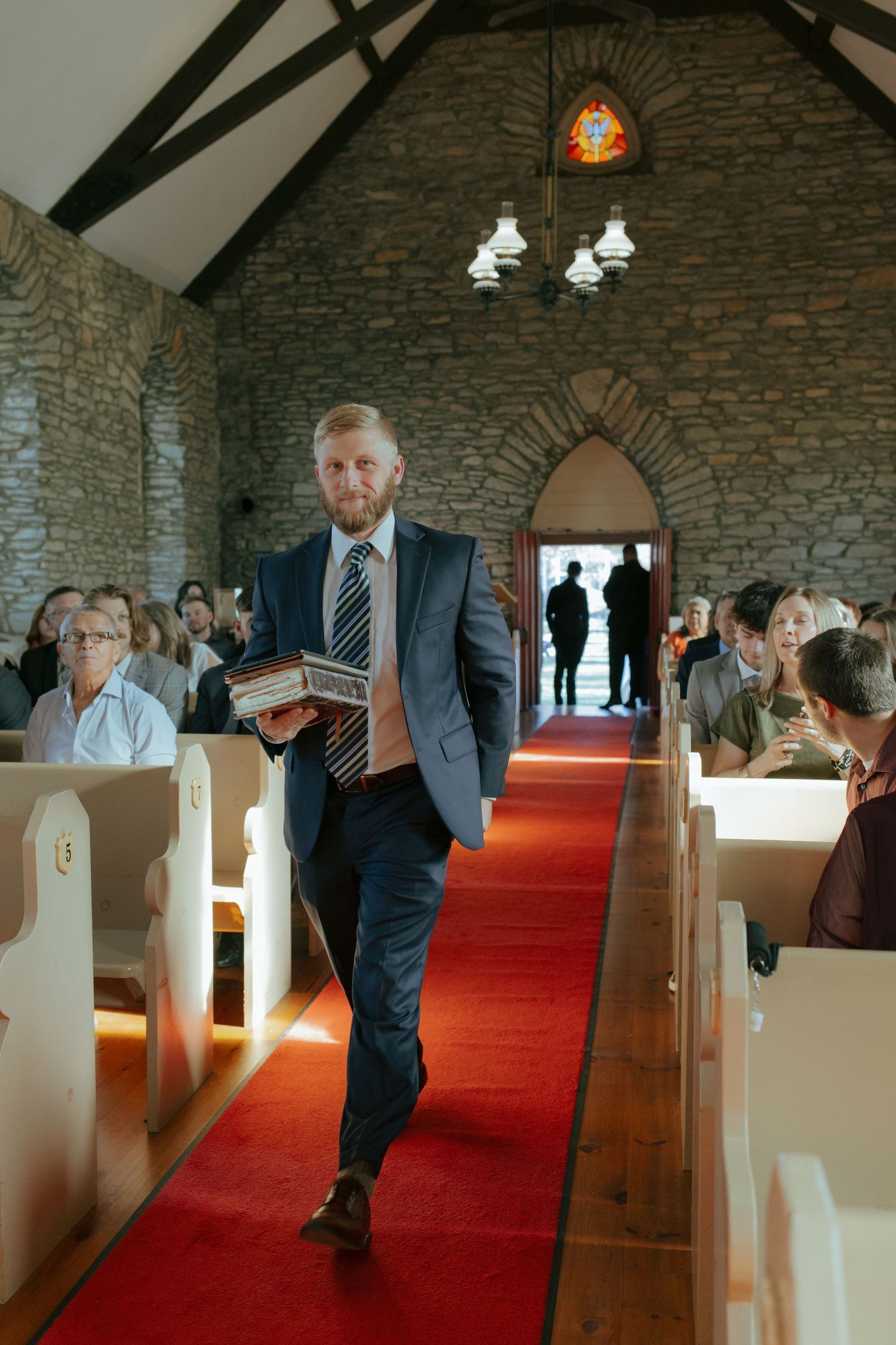 Man in suit walking down red carpet aisle in stone church during wedding ceremony.