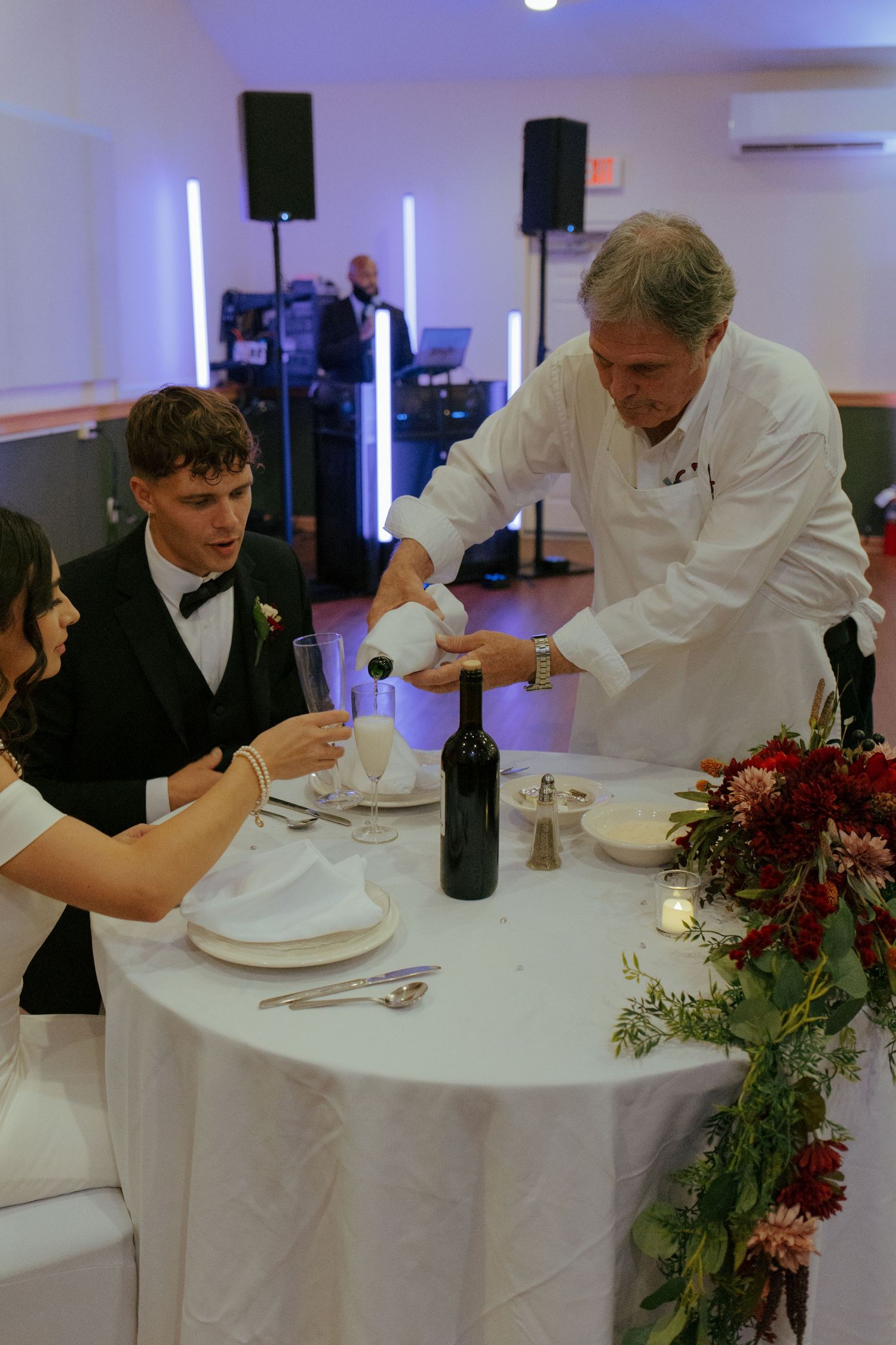 Chef pours champagne for newlywed couple seated at elegant white reception table with floral centerpiece.