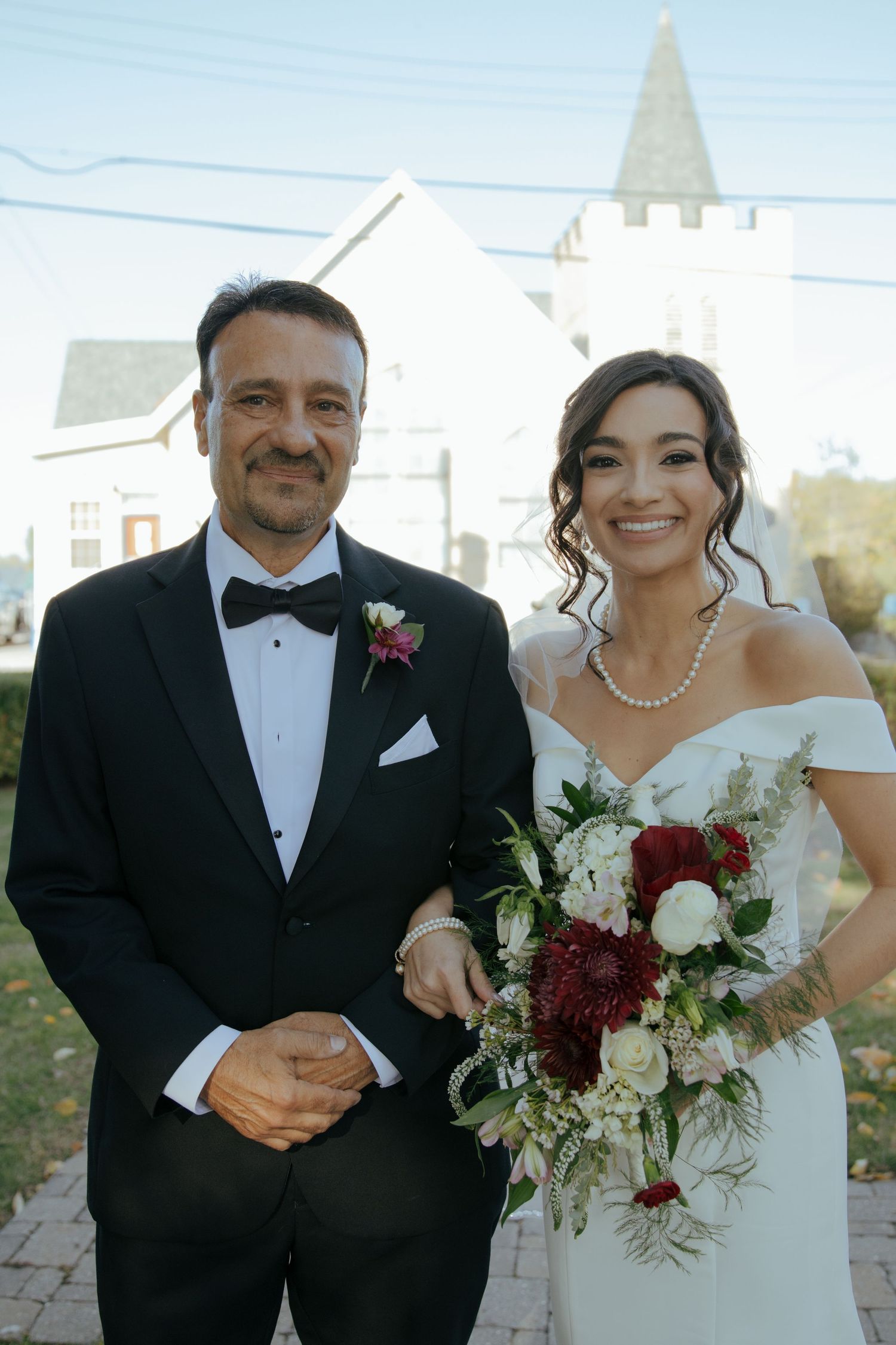 Smiling bride holding a bouquet of red and white flowers poses with a man in a tuxedo outside.