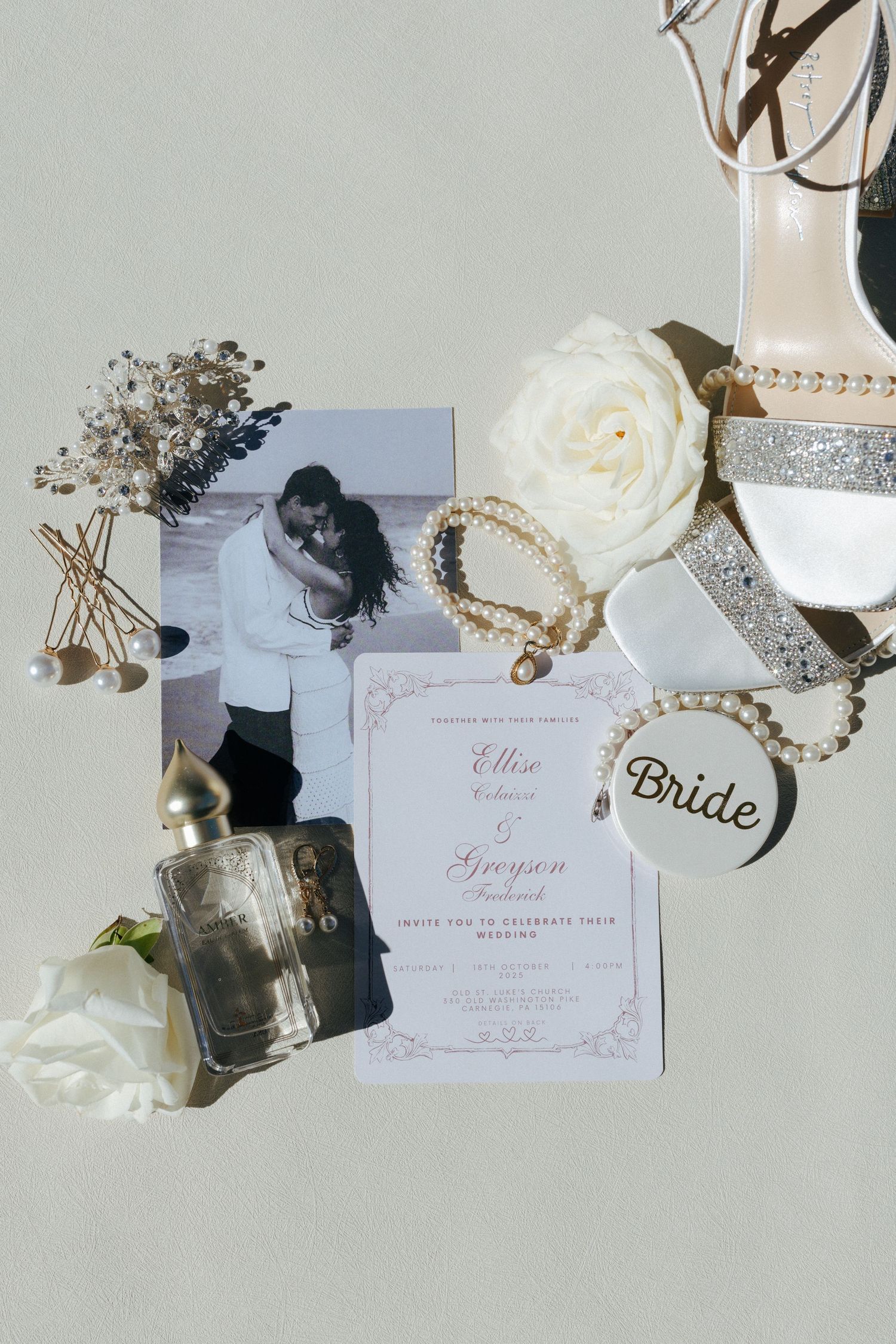 Elegant wedding flat lay featuring invitations, pearl jewelry, glitter heels, white roses, perfume, and a bride tag on a neutral background.