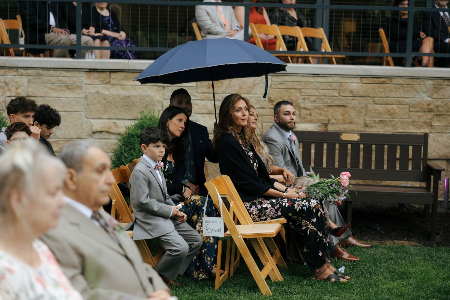 Wedding guests seated outdoors, one holding black umbrella, at garden ceremony with stone wall backdrop.