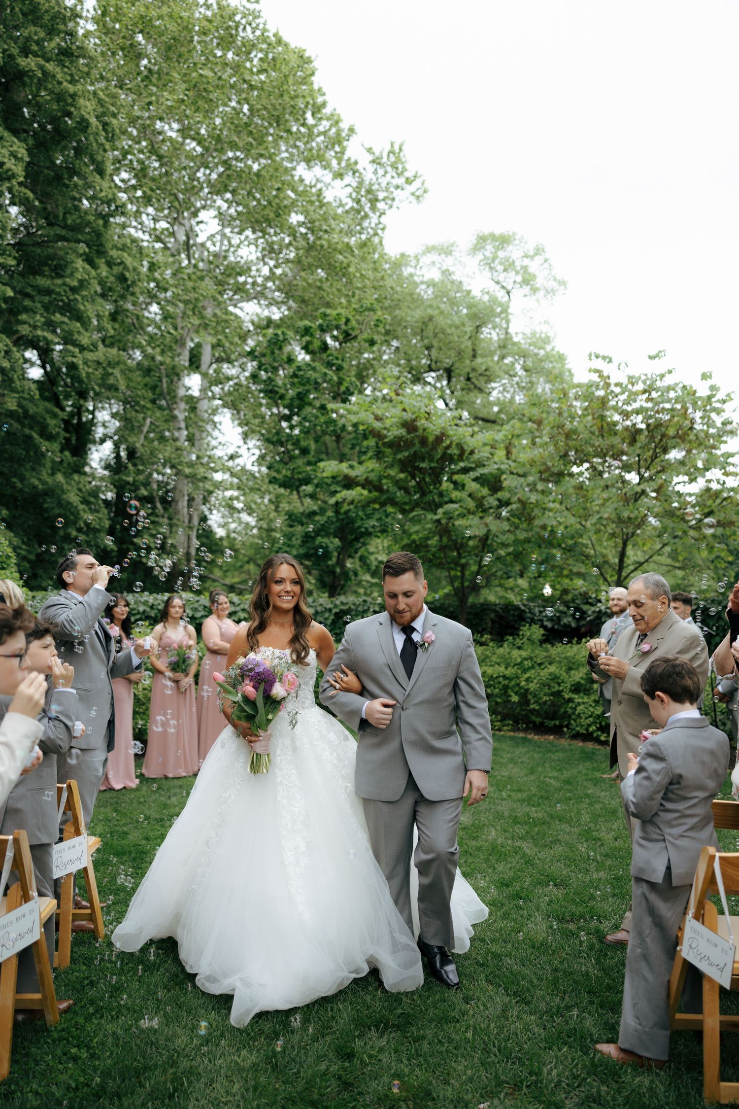 Newlyweds walk back down aisle after outdoor garden wedding ceremony as guests celebrate around them.