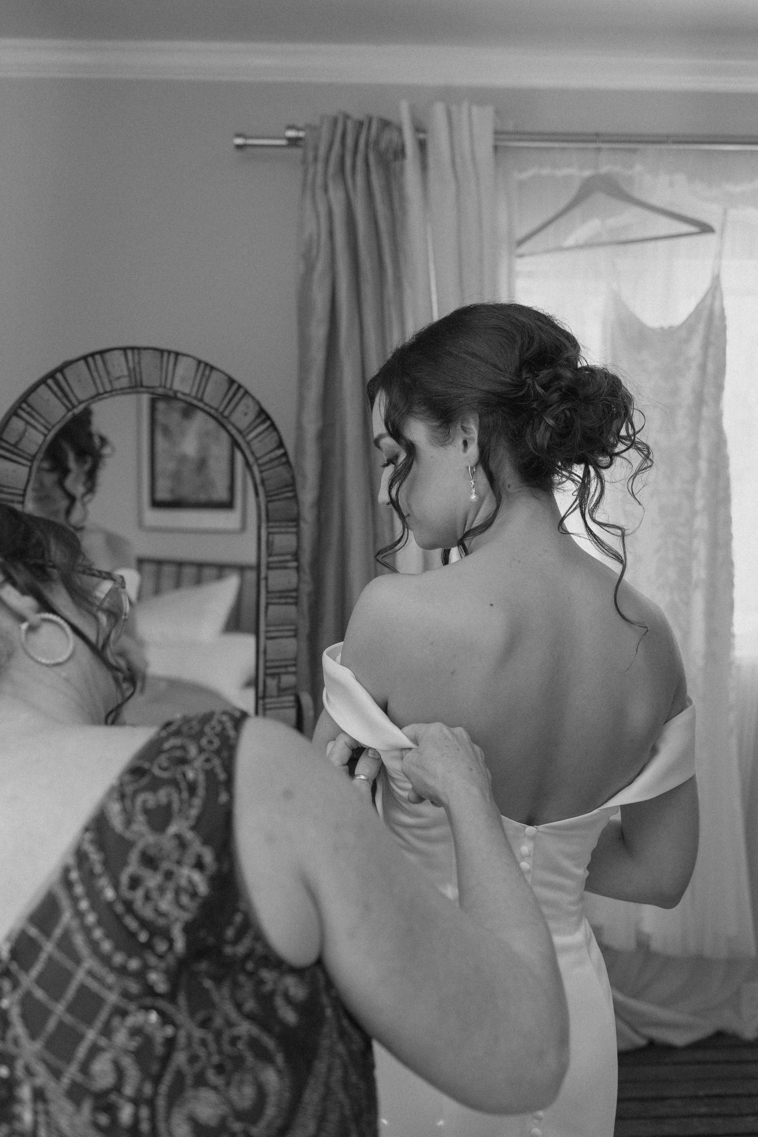 Black and white photo of bride in off-shoulder wedding dress being helped by assistant near mirror.