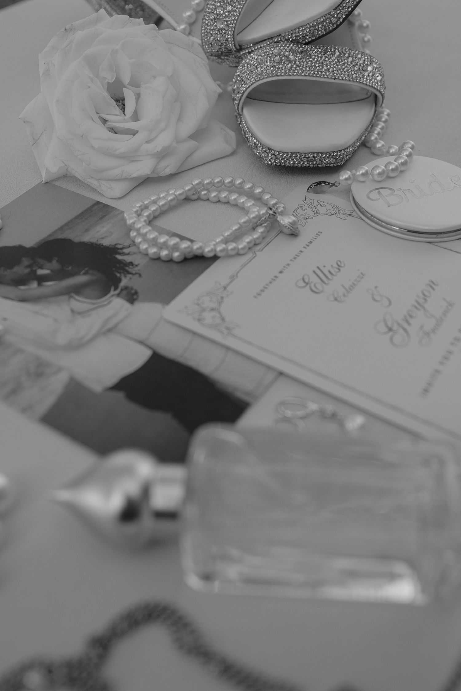 Black and white flat lay of wedding accessories including pearl bracelet, rhinestone heels, rose, and elegant stationery.
