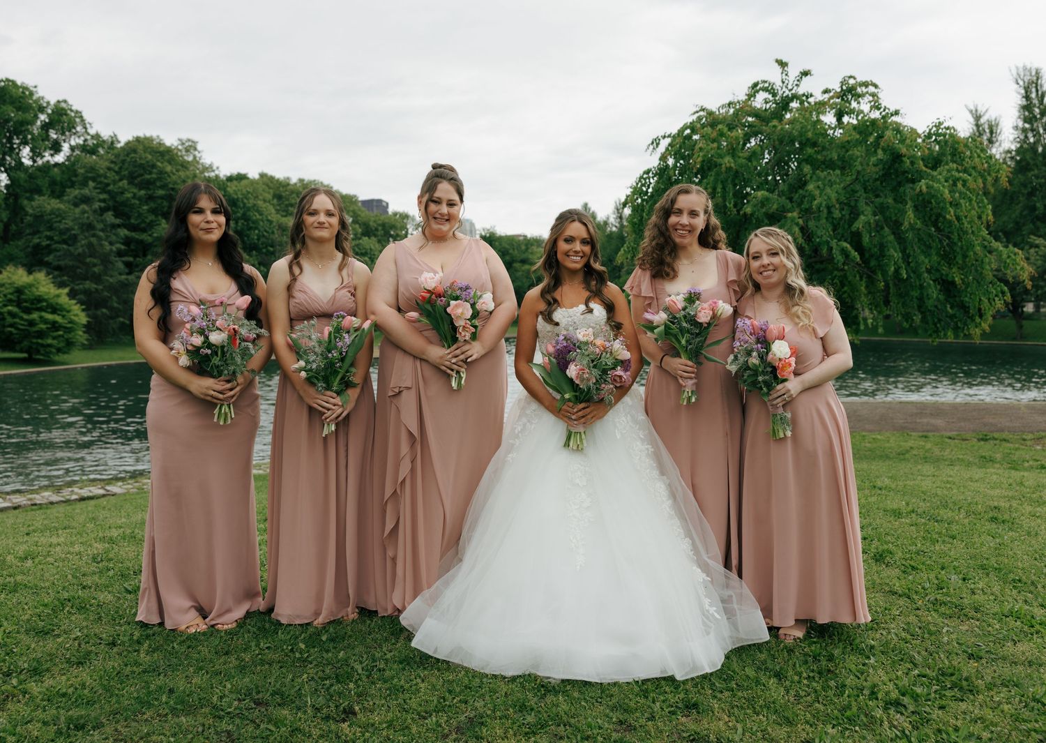 Bride and four bridesmaids in dusty rose gowns pose with bouquets by a scenic waterfront.