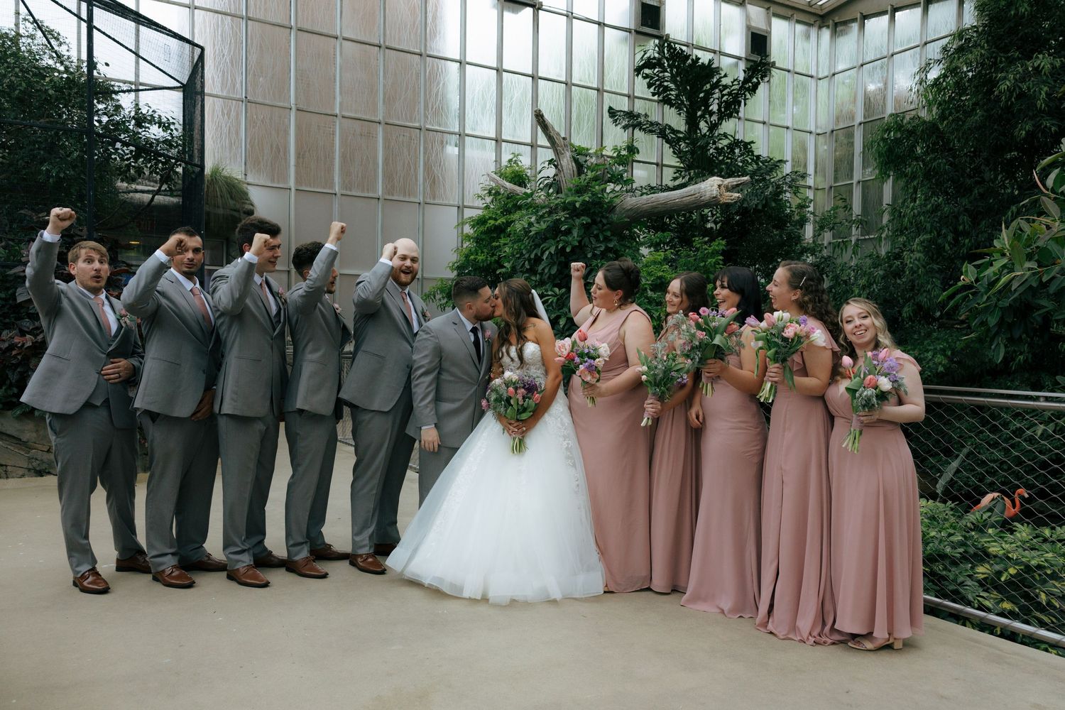 Wedding party celebrating outdoors, groomsmen in gray suits and bridesmaids in dusty rose gowns, with bride and groom at center.