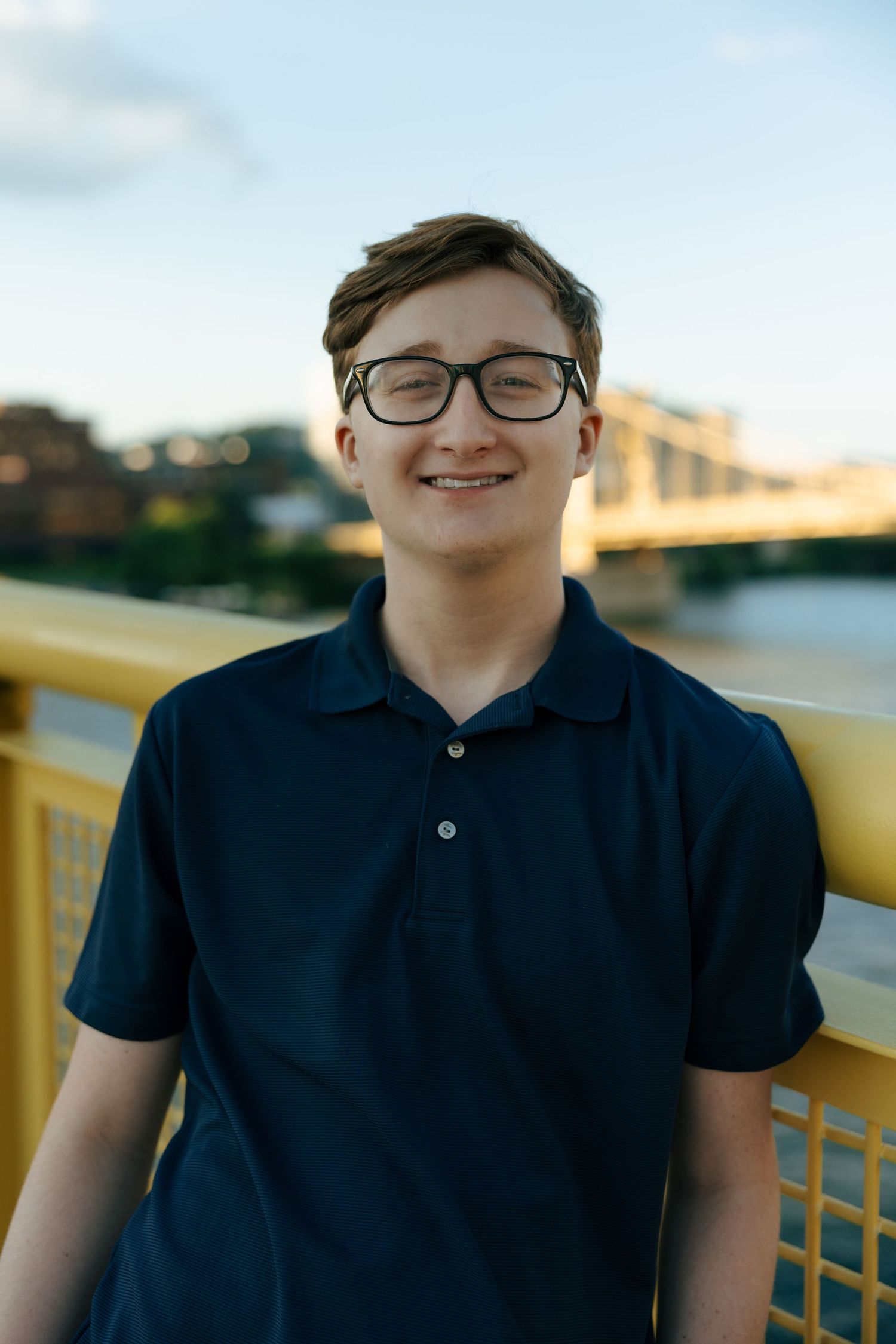 Smiling teenage boy with glasses in navy polo shirt standing on yellow bridge at sunset.